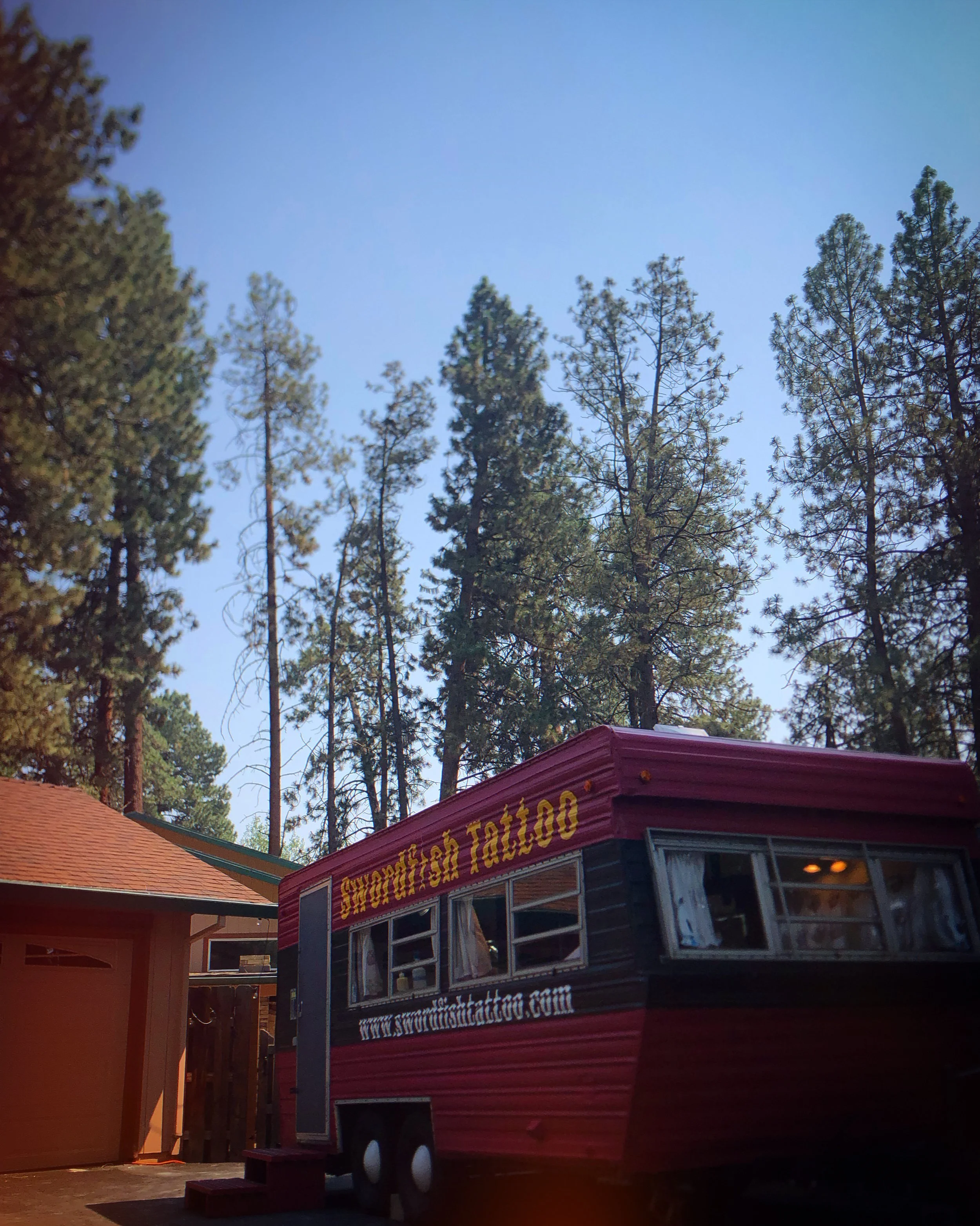 A red food truck with yellow lettering, parked outdoors among tall pine trees with a partly cloudy sky above.