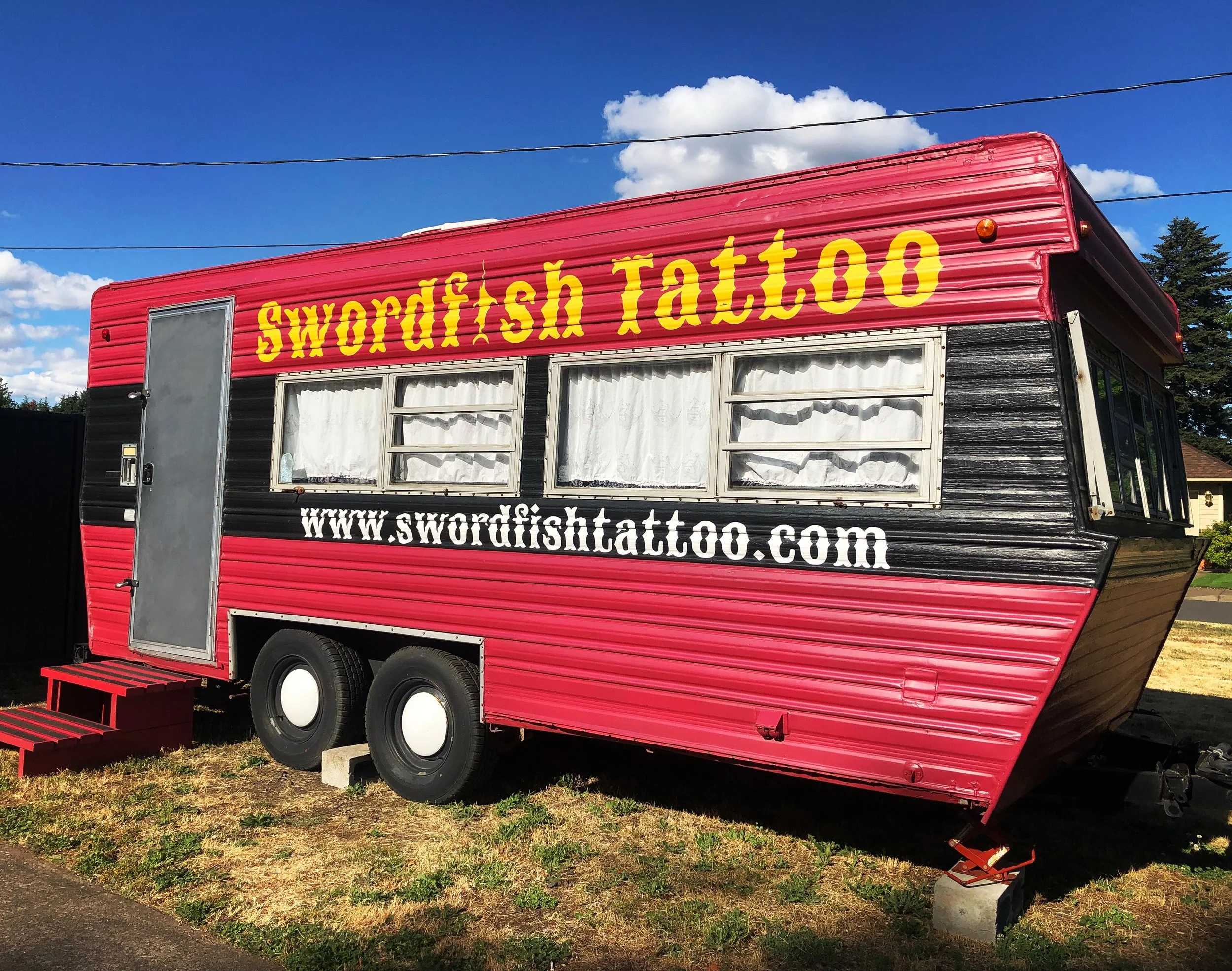 A red food truck with the words "Swordfish Tattoo" painted in yellow on the side. The truck has black and white accents, and windows covered with white curtains. There are red steps leading up to the entrance. The truck is parked on grass with blue s