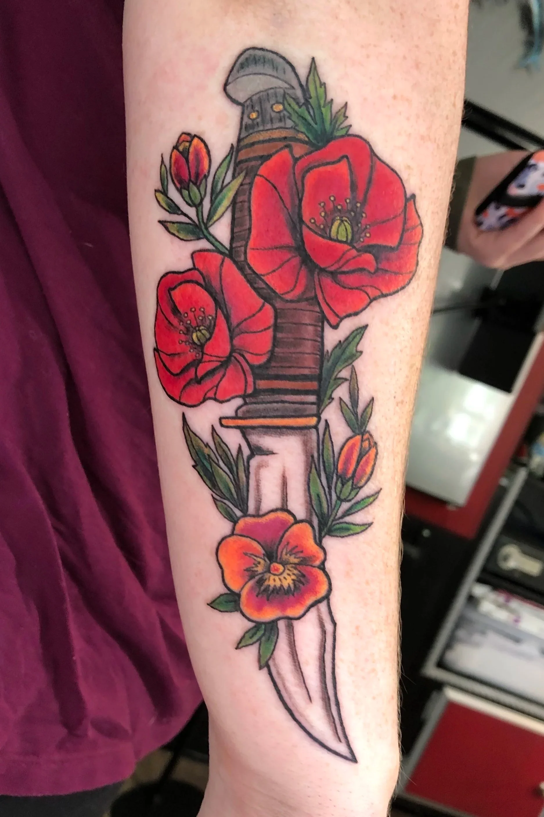 A colorful tattoo of red poppy flowers with green leaves and a black and gray dagger on a person's arm.