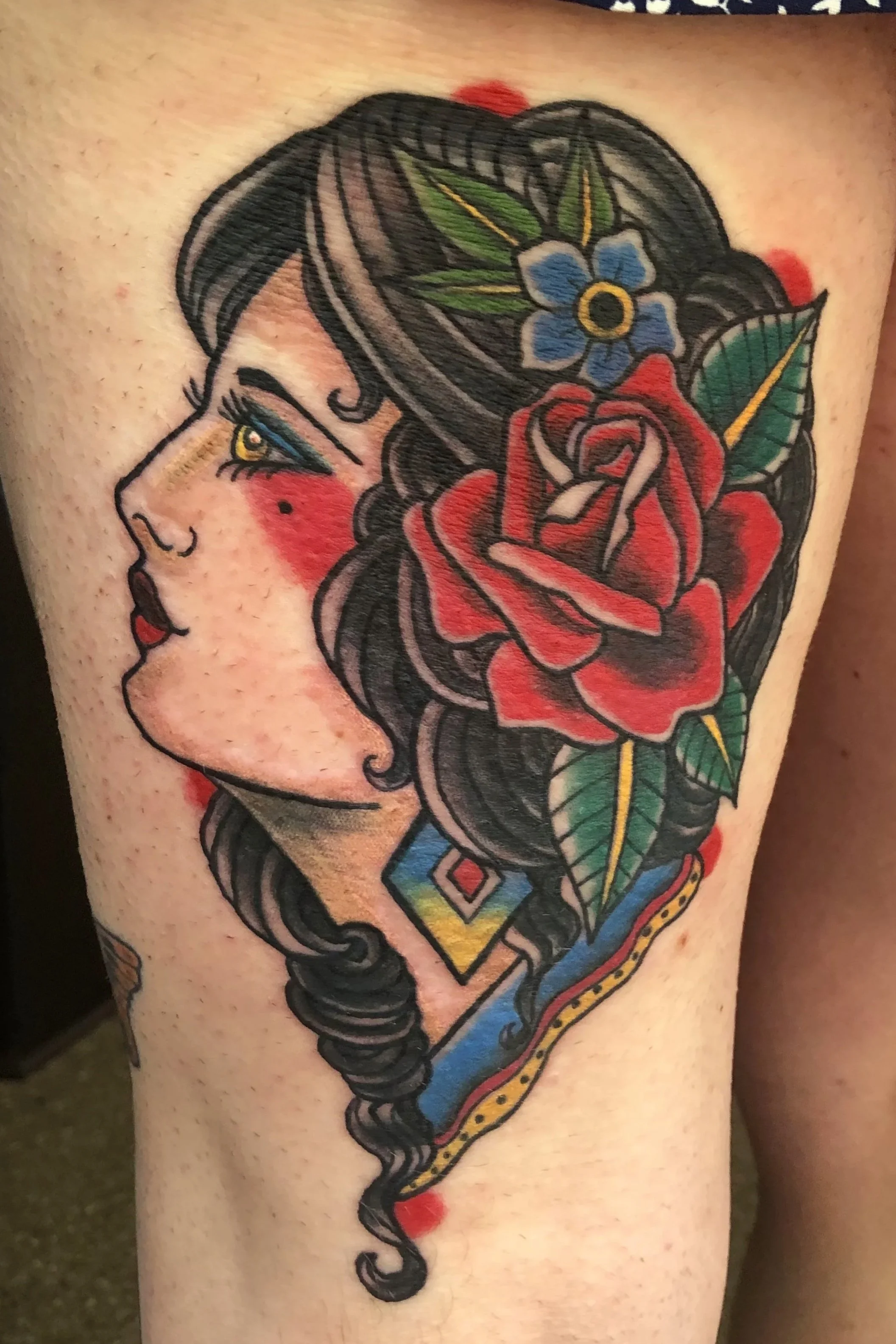 Tattoo of a woman with dark hair decorated with a blue flower and green leaves, red roses, and geometric patterns.