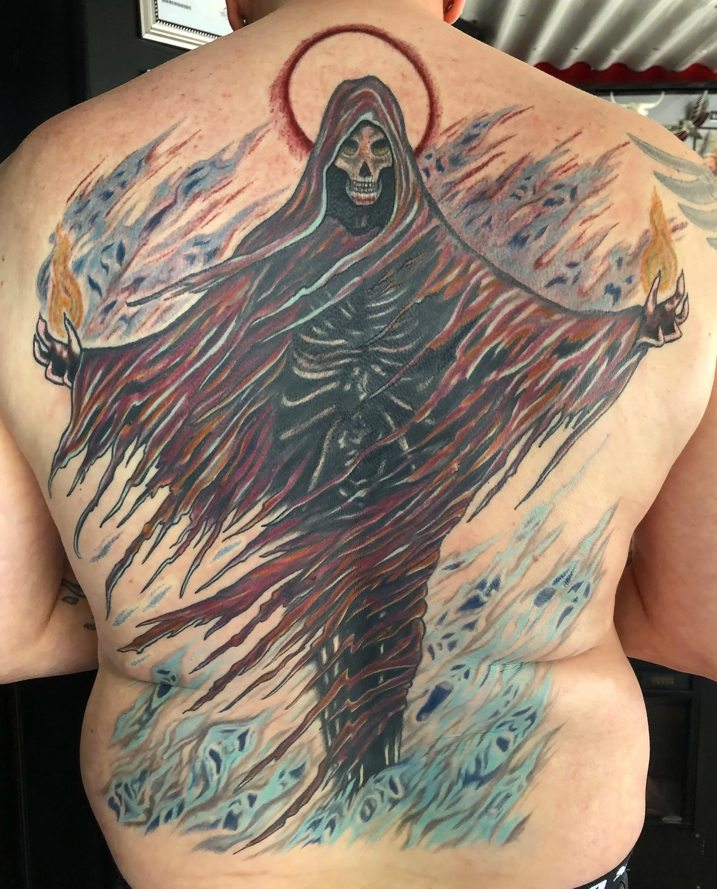 Tattoo of a skull-headed grim reaper figure with a dark hood and outstretched arms, surrounded by flames and abstract designs, on a person's back.