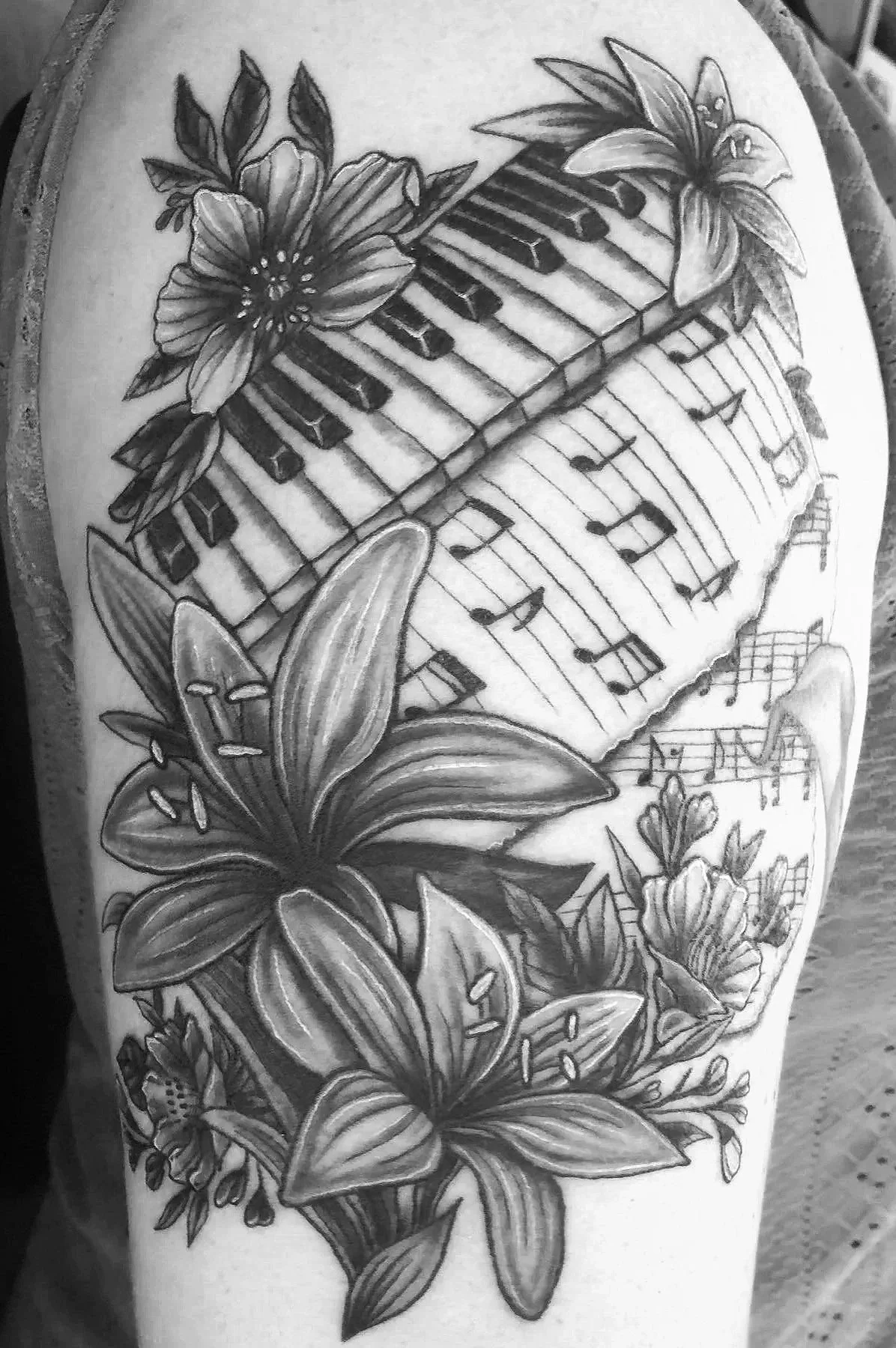 Black and gray tattoo of flowers and leaves surrounding a musical staff with notes and a piano keyboard overlay.