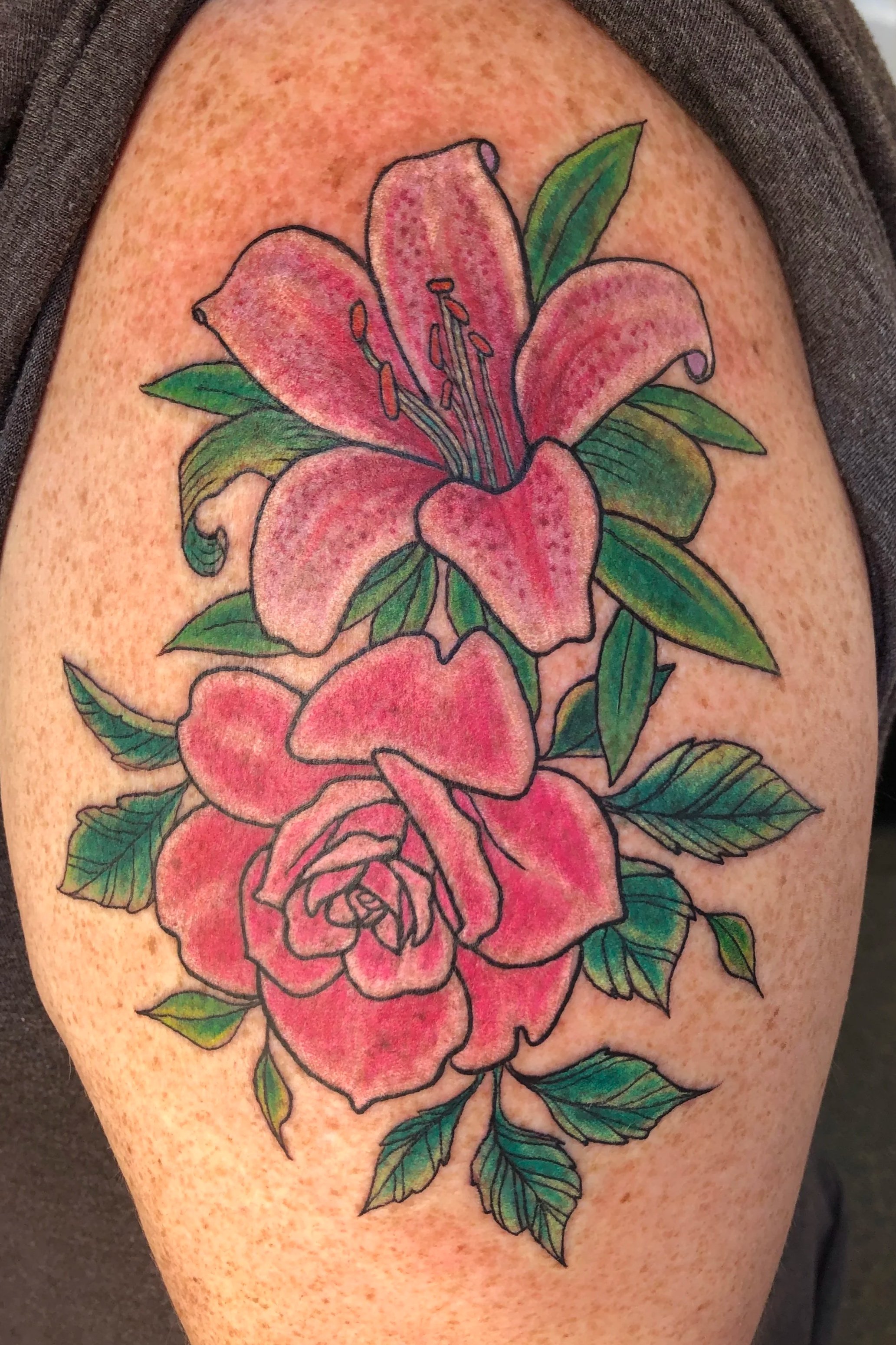 Colorful tattoo of a pink lily flower with green leaves on a person's upper arm.