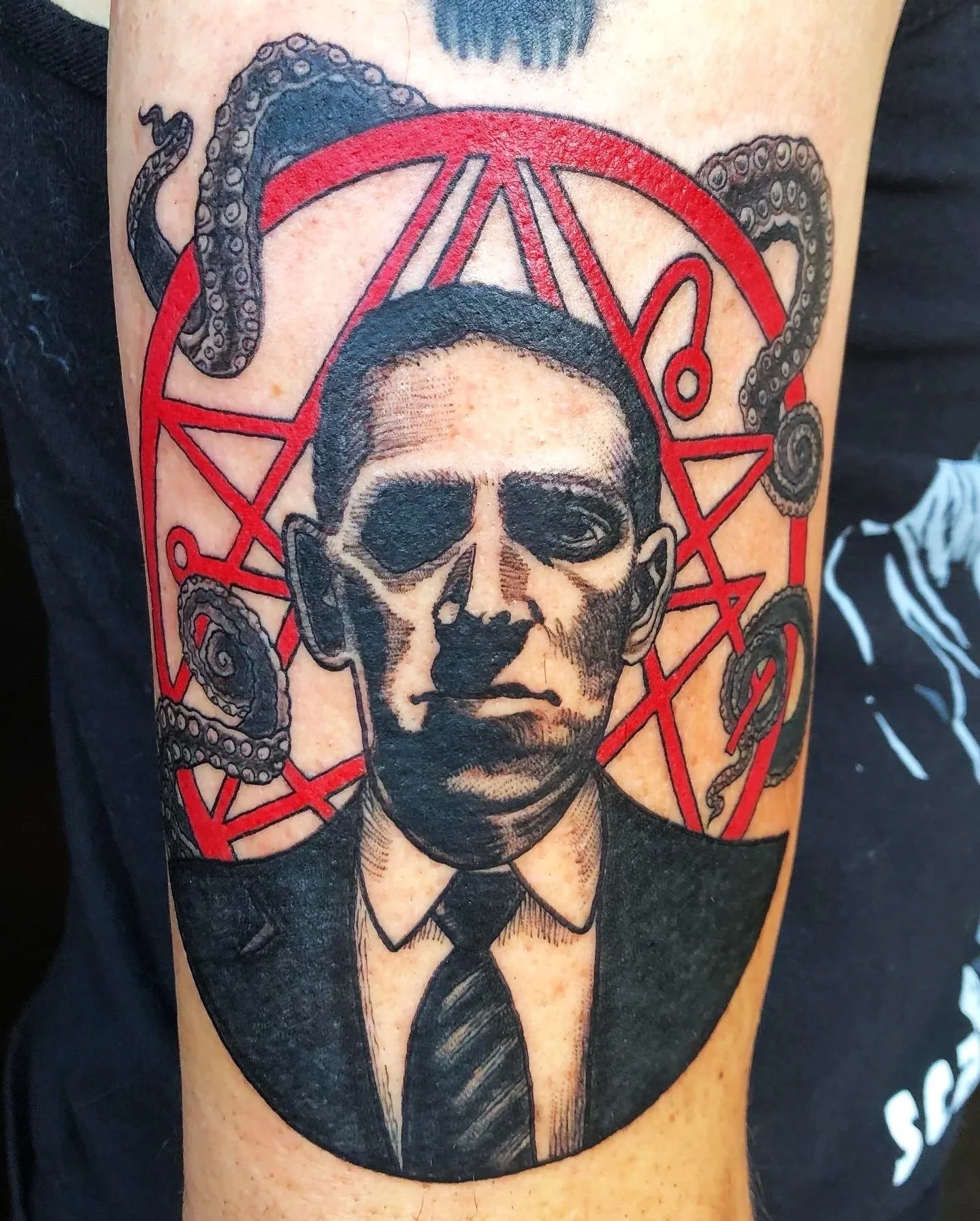 Tattoo of a black and gray portrait of a man with short hair, wearing a formal shirt and tie, with a red pentagram symbol and coiled octopus tentacles surrounding his head.