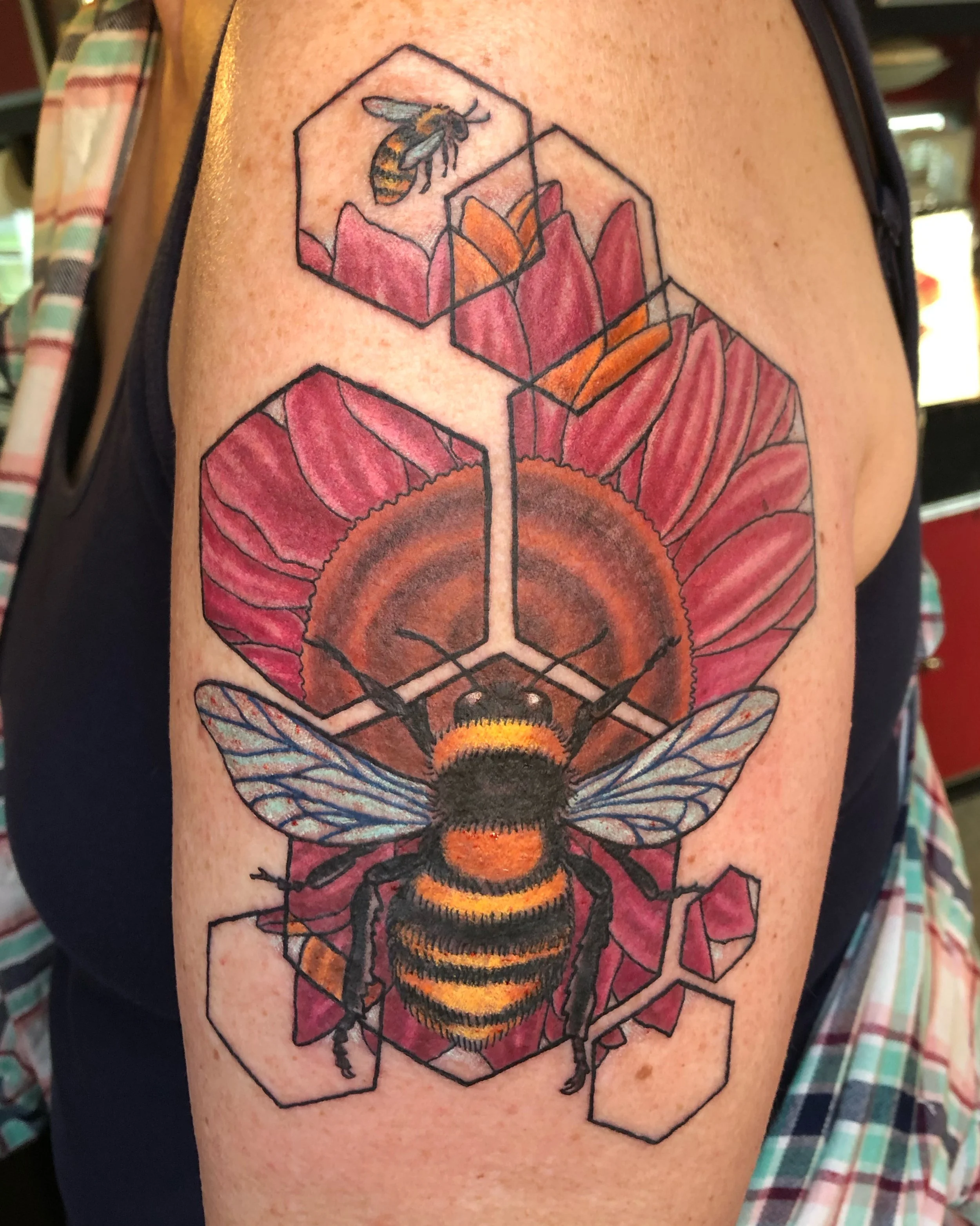 Colorful tattoo of a bee, within a geometric honeycomb design, on a person's upper arm.