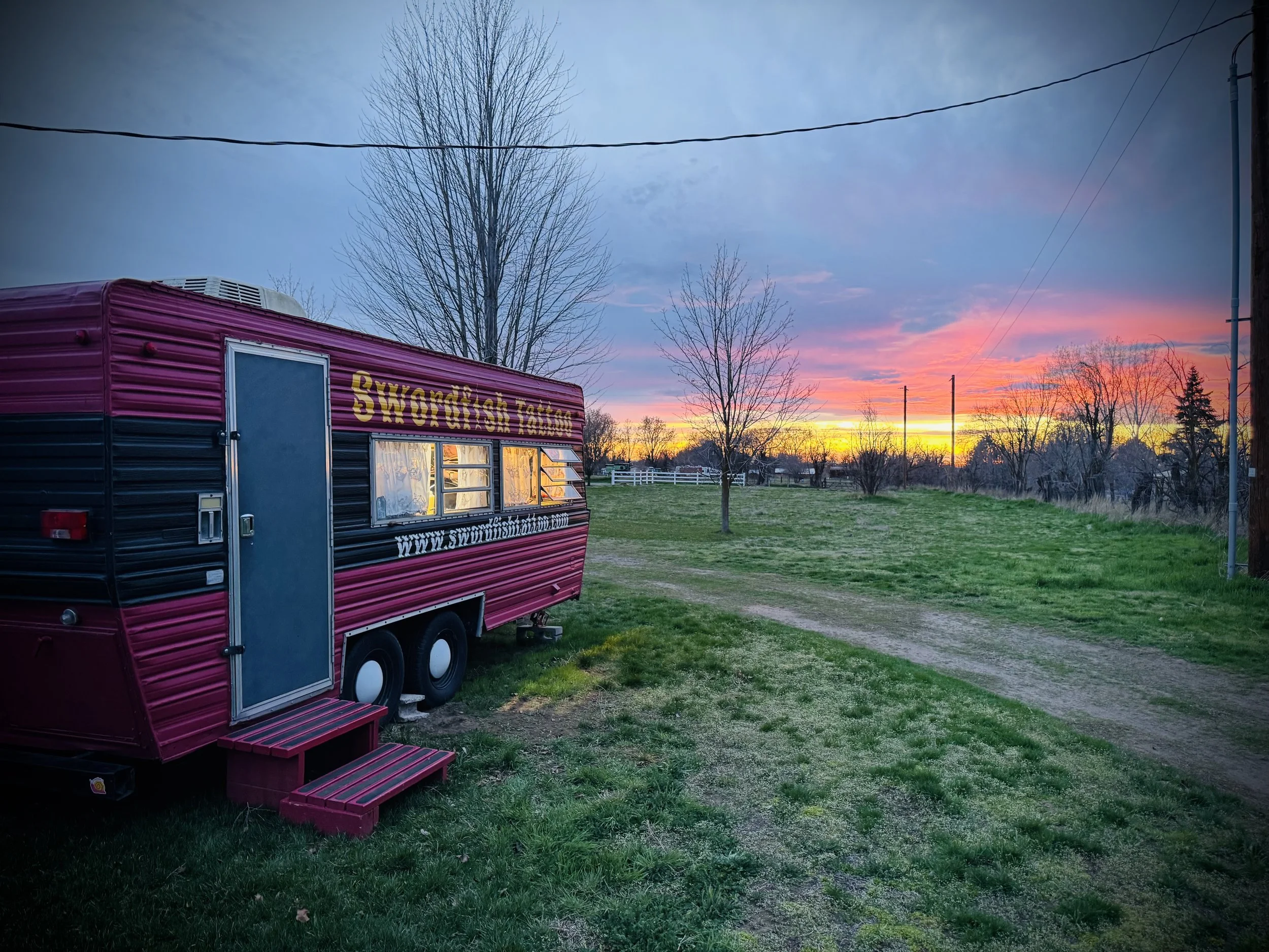 A red food truck with yellow text that reads "Swordfish Tattoo" parked on a grassy area during sunset, with bare trees, power lines, and a multicolored sky in the background.
