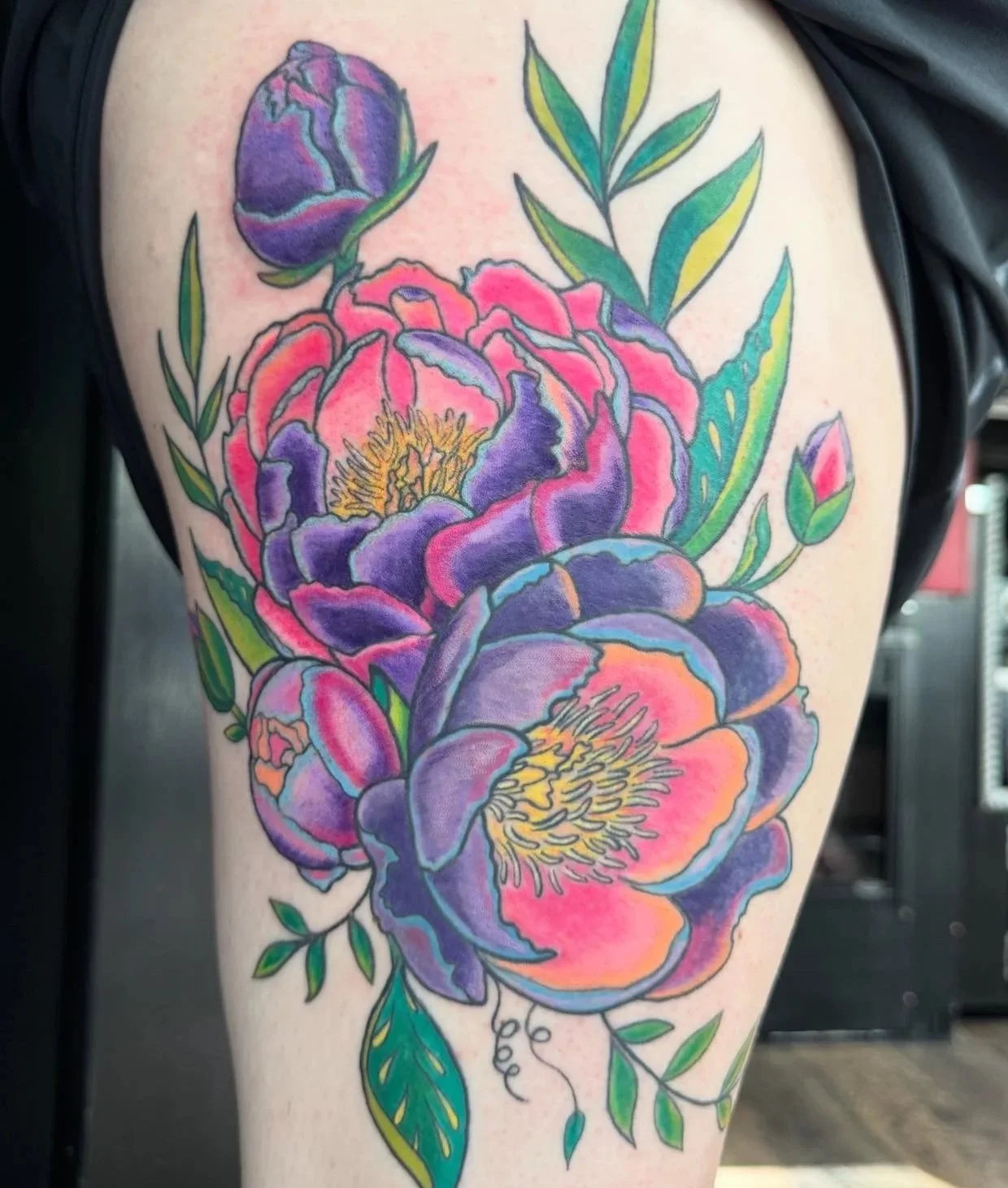 Colorful floral tattoo of purple, pink, and orange peonies with green leaves on a person's thigh.