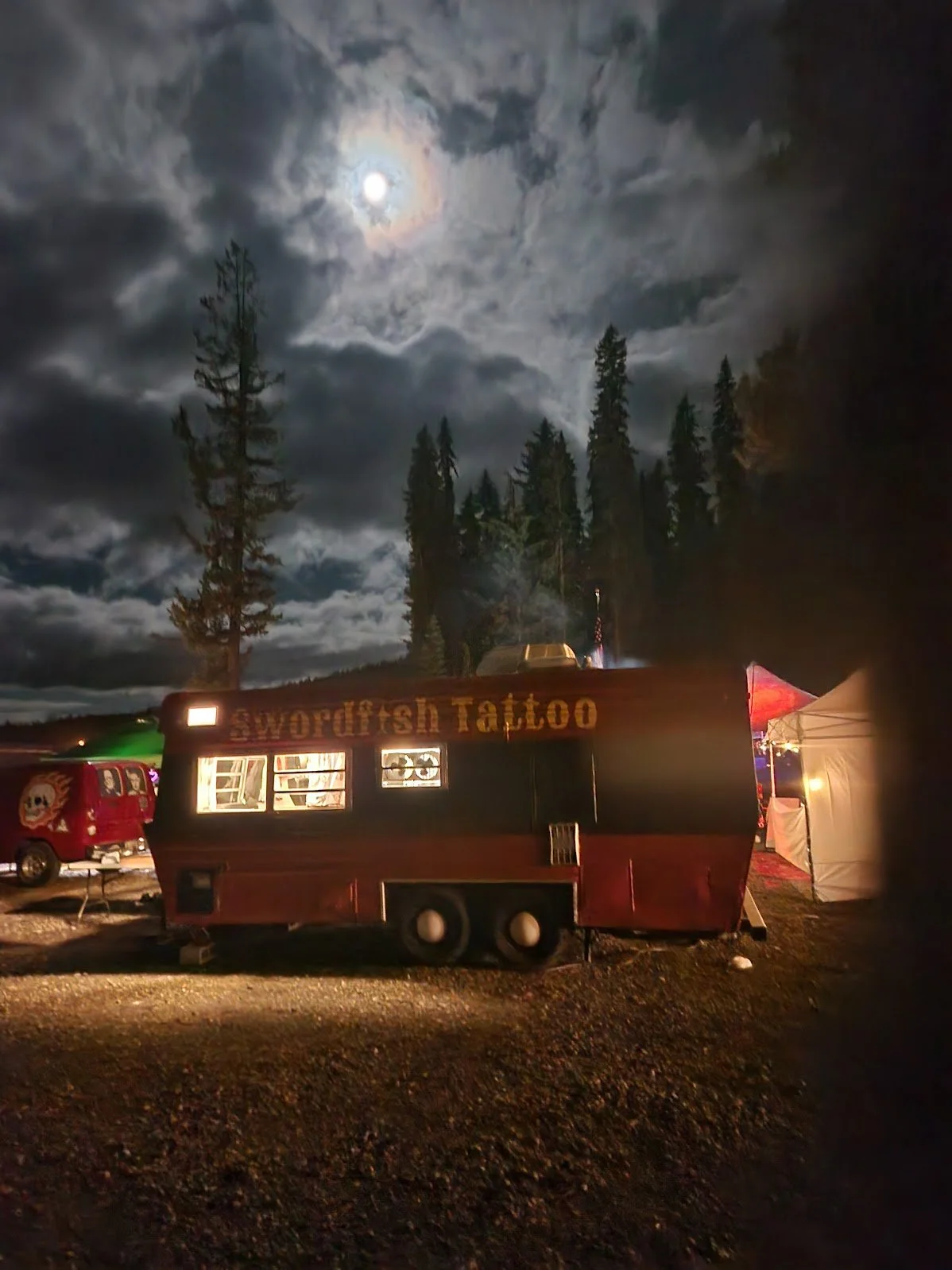 Night scene with a full moon behind clouds, tall trees, and a red trailer that reads 'Swordfish Tattoo' parked on gravel with tents and other vehicles nearby.