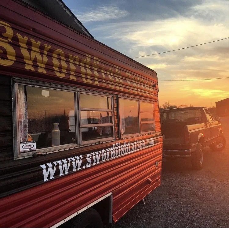 A red and black food trailer with a website URL on the side, parked next to a pickup truck during sunset.