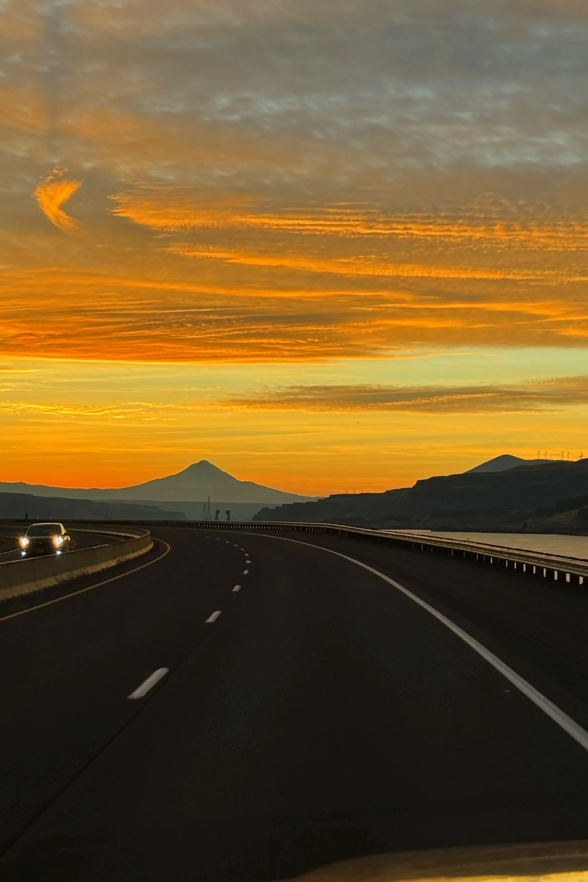 A winding highway curves through a landscape at sunset, with mountains in the distance and a sky filled with layered orange, yellow, and gray clouds.