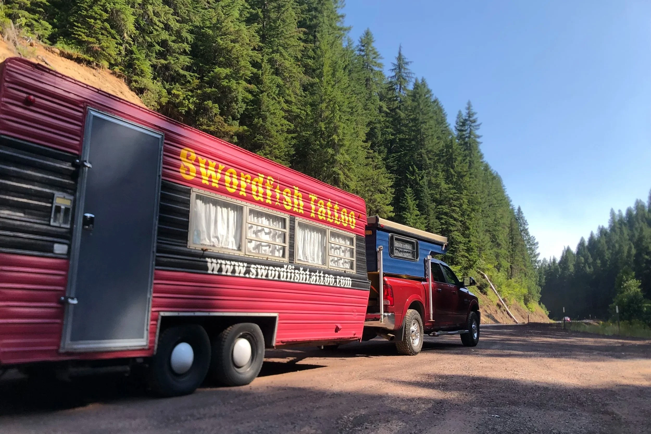 A red mobile tattoo studio with yellow lettering that reads 'Swordfish Tattoo' parked next to a black pickup truck on a paved road, with tall green pine trees and a blue sky in the background.