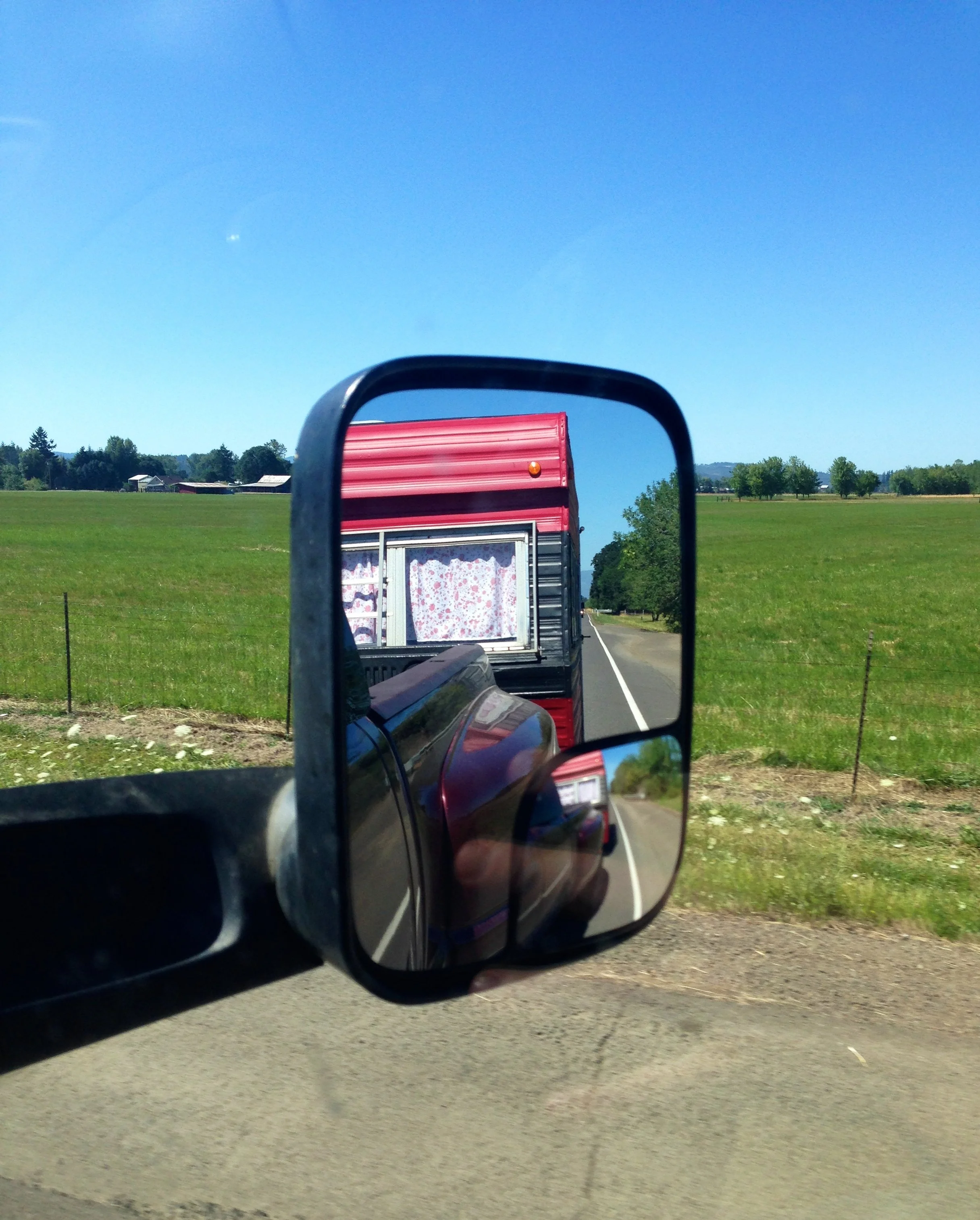 A side mirror of a vehicle reflecting the back of a red trailer with floral curtains, on a rural road with green fields and trees, under a clear blue sky.