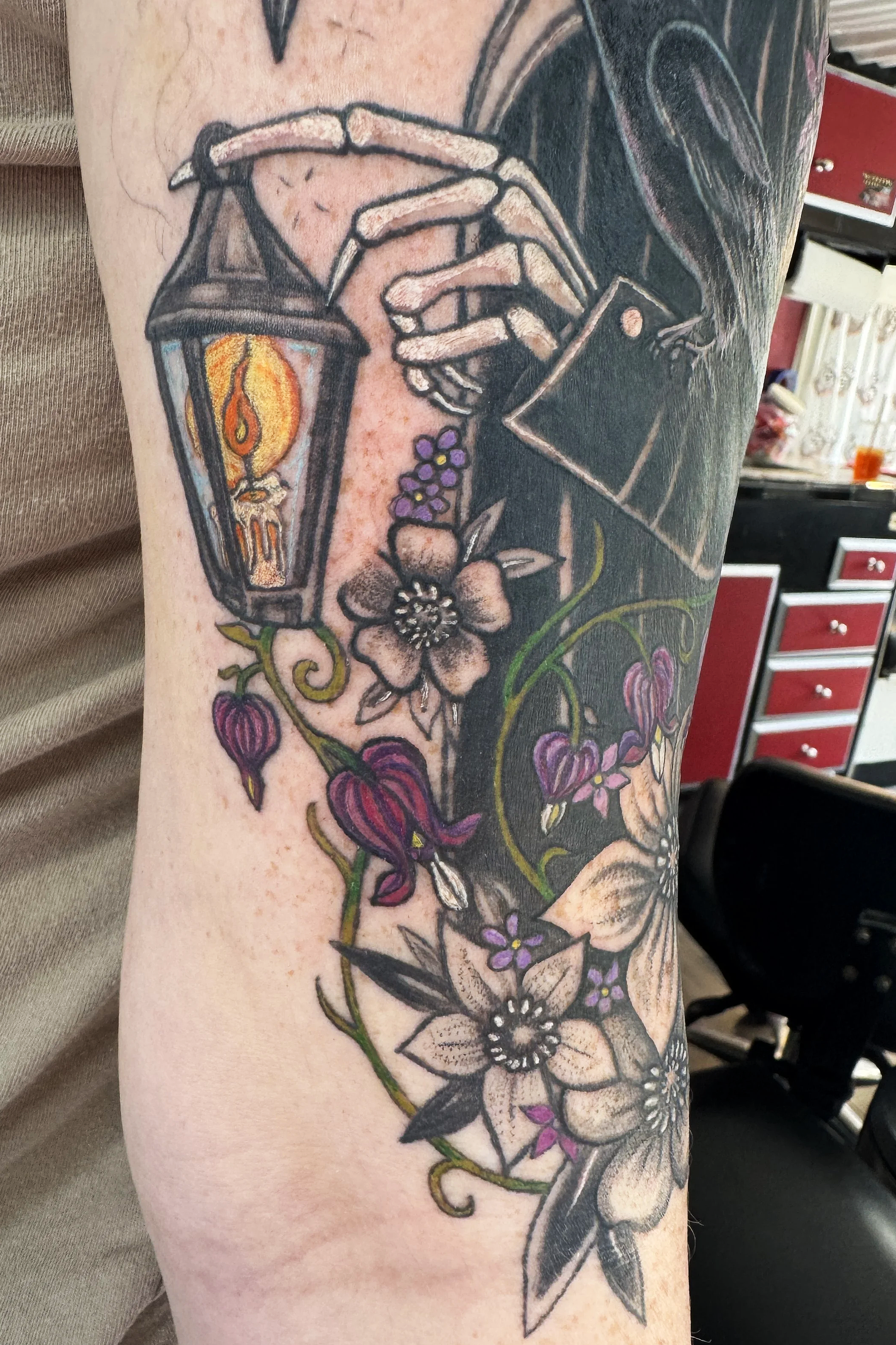 Tattoo of a lantern with a candle, surrounded by various flowers and leaves on a person's arm.