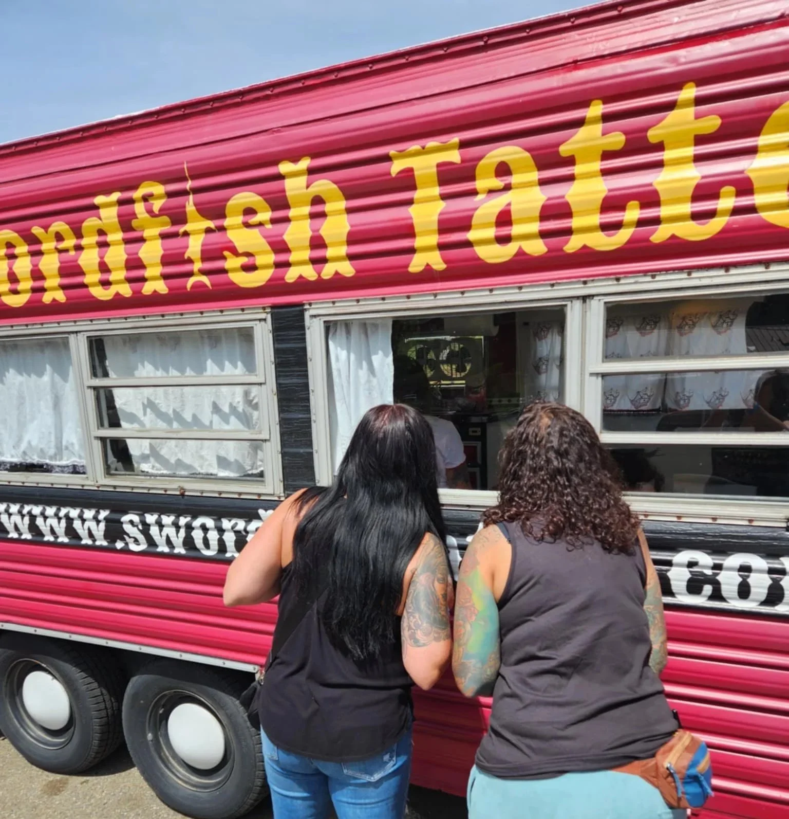 Two women with tattoos are lining up at a fmobile tattoo studio with a red and black exterior. The mobile tattoo studio has a sign with yellow text and is parked outdoors on a sunny day.
