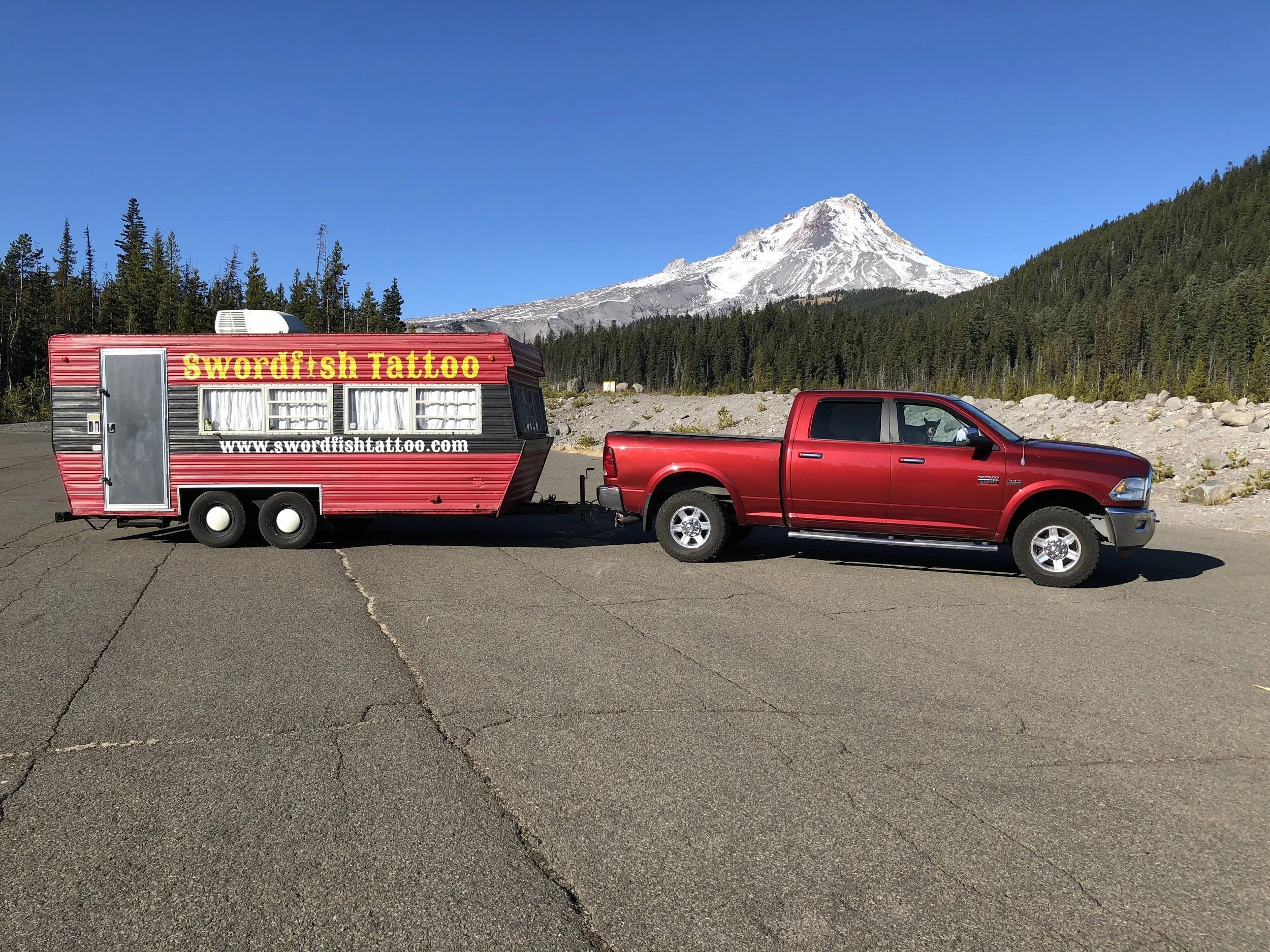 A red truck with a travel trailer attached parked on a paved surface with cracks, with a backdrop of green forested hills and Mount Hood snowy peak under a clear blue sky.