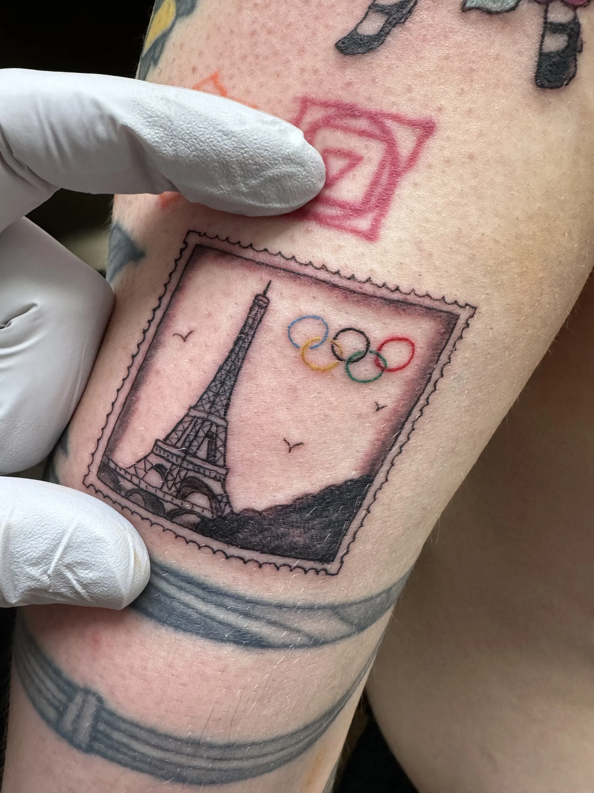 A tattoo of the Eiffel Tower with the Olympic rings underneath, surrounded by a postage stamp border on a person's skin.