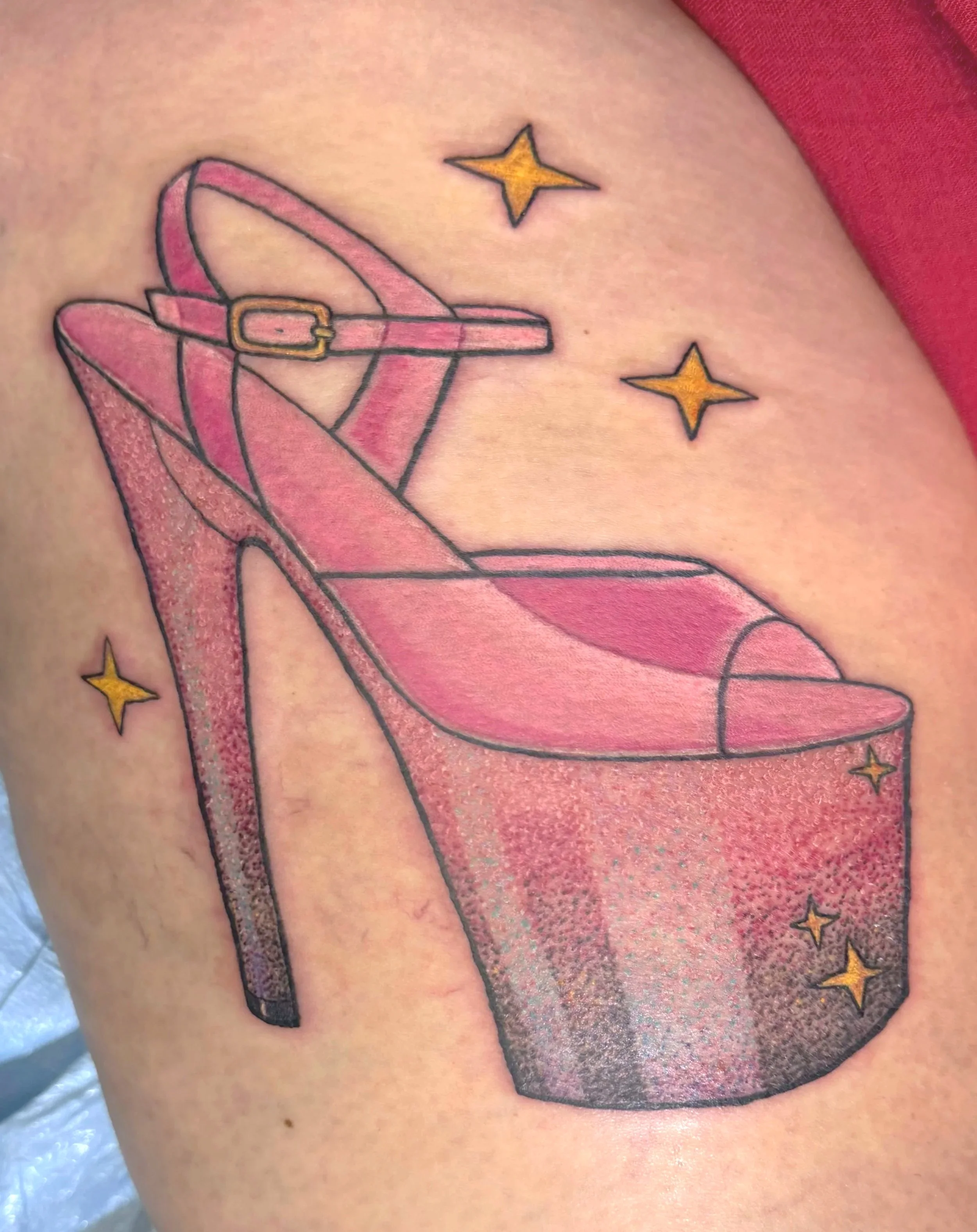 Tattoo of a pink high-heeled shoe with a strap and a gold buckle, decorated with yellow stars, on a person's skin.