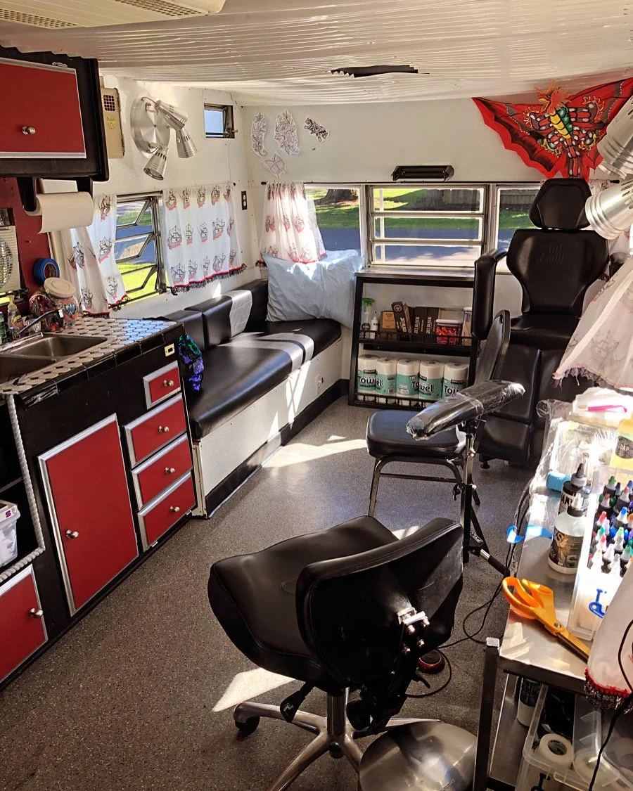 Inside a mobile tattoo studio with black and red cabinetry, tattoo chairs, and supplies, illuminated by natural light from windows, with decorations and artwork on the walls.
