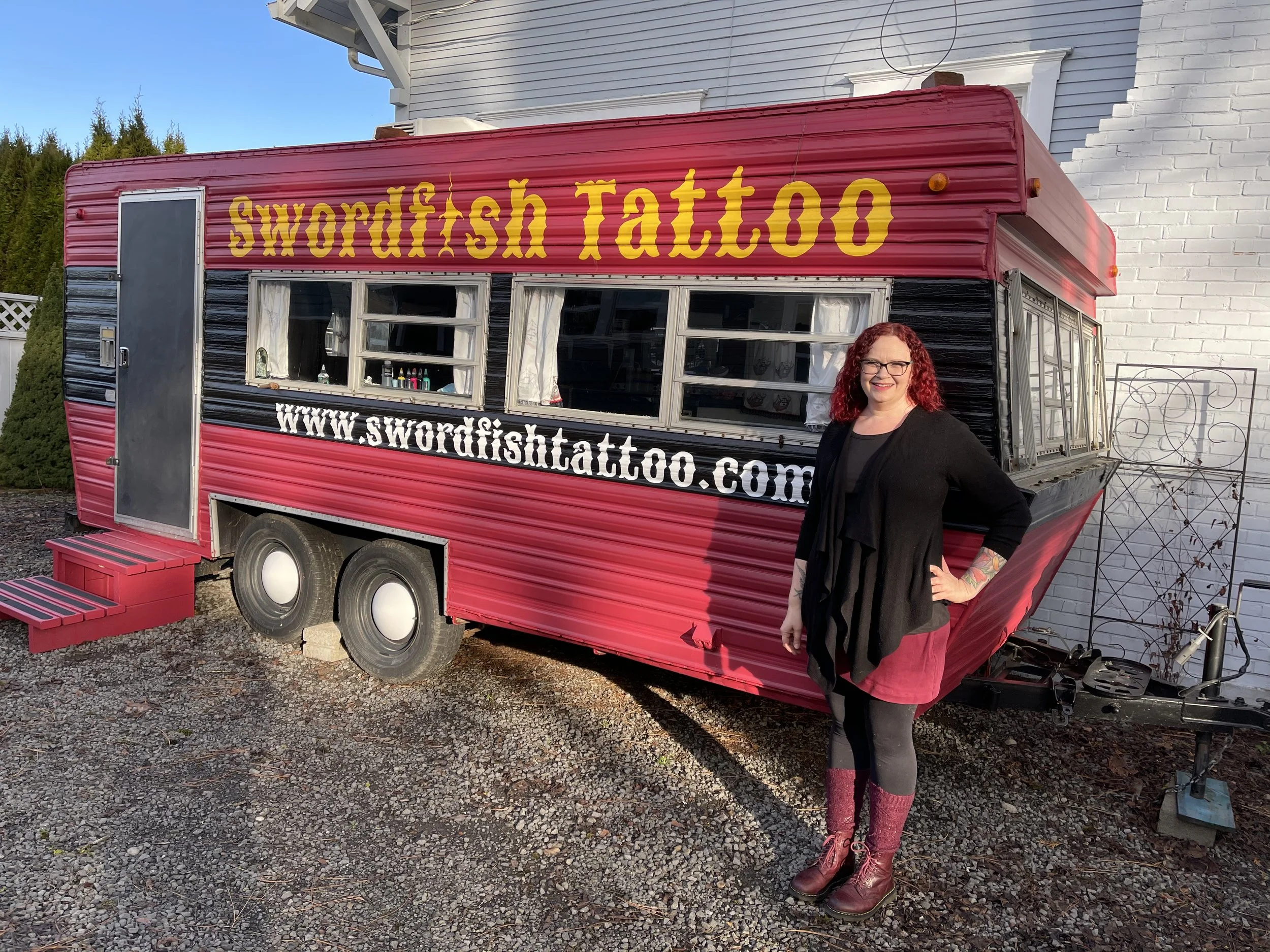 A red food trailer with the words "Swordfish Tattoo" written on it, and a woman with red hair, glasses, black clothing, and tan boots standing next to it, smiling.