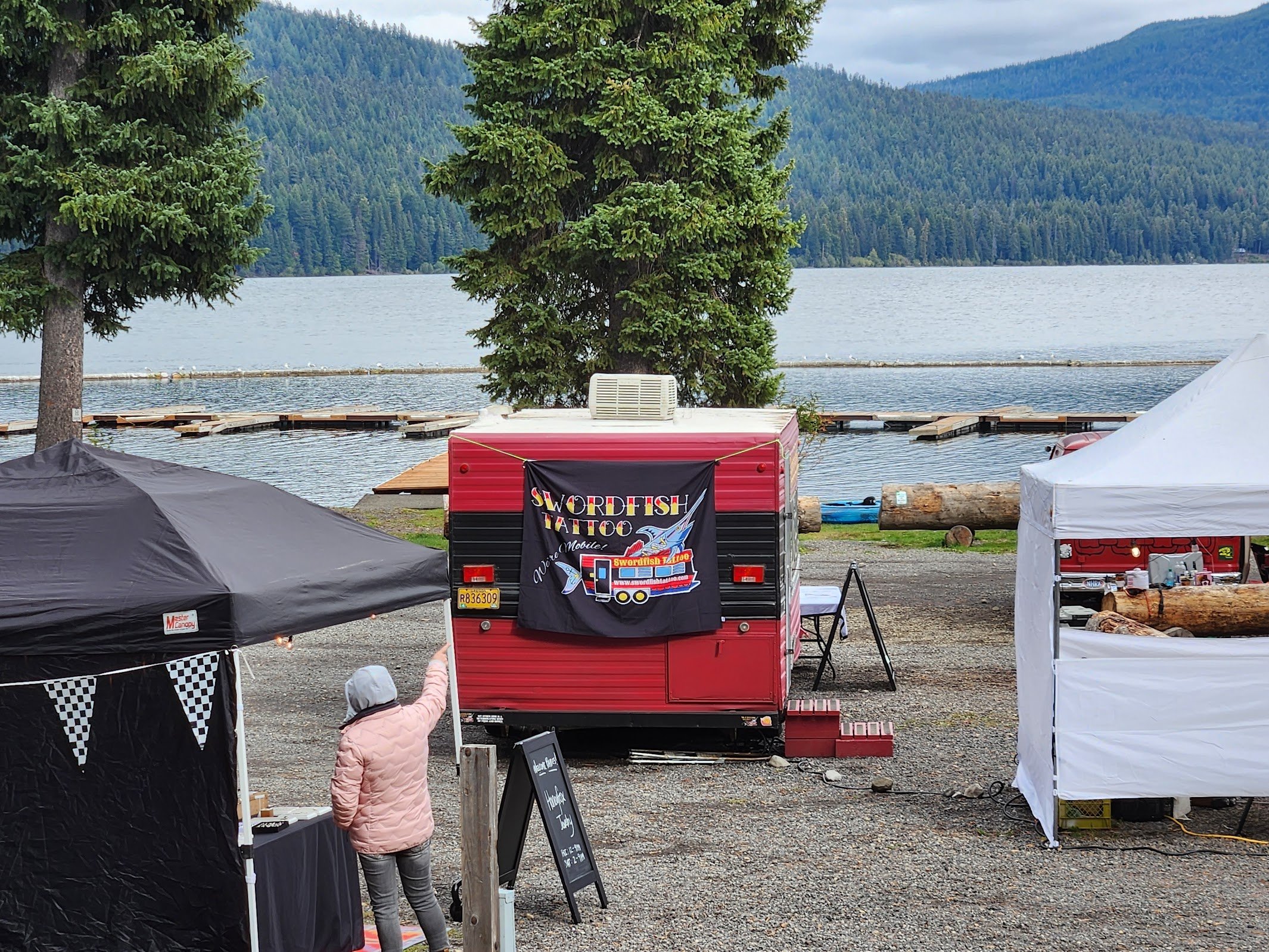 Outdoor marketplace by a lake with trees and mountains in the background, featuring tents, a food trailer with a banner reading 'Swordfish Tattoo', and a person in a pink jacket and white hat.
