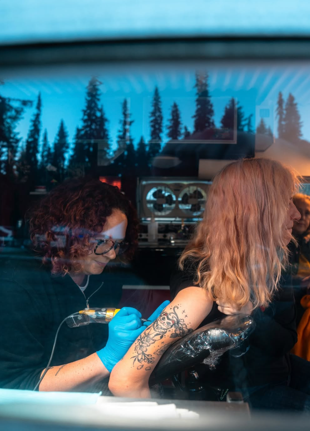 A woman is getting a tattoo on her upper arm by a tattoo artist, who is wearing glasses and blue gloves, inside a vehicle.