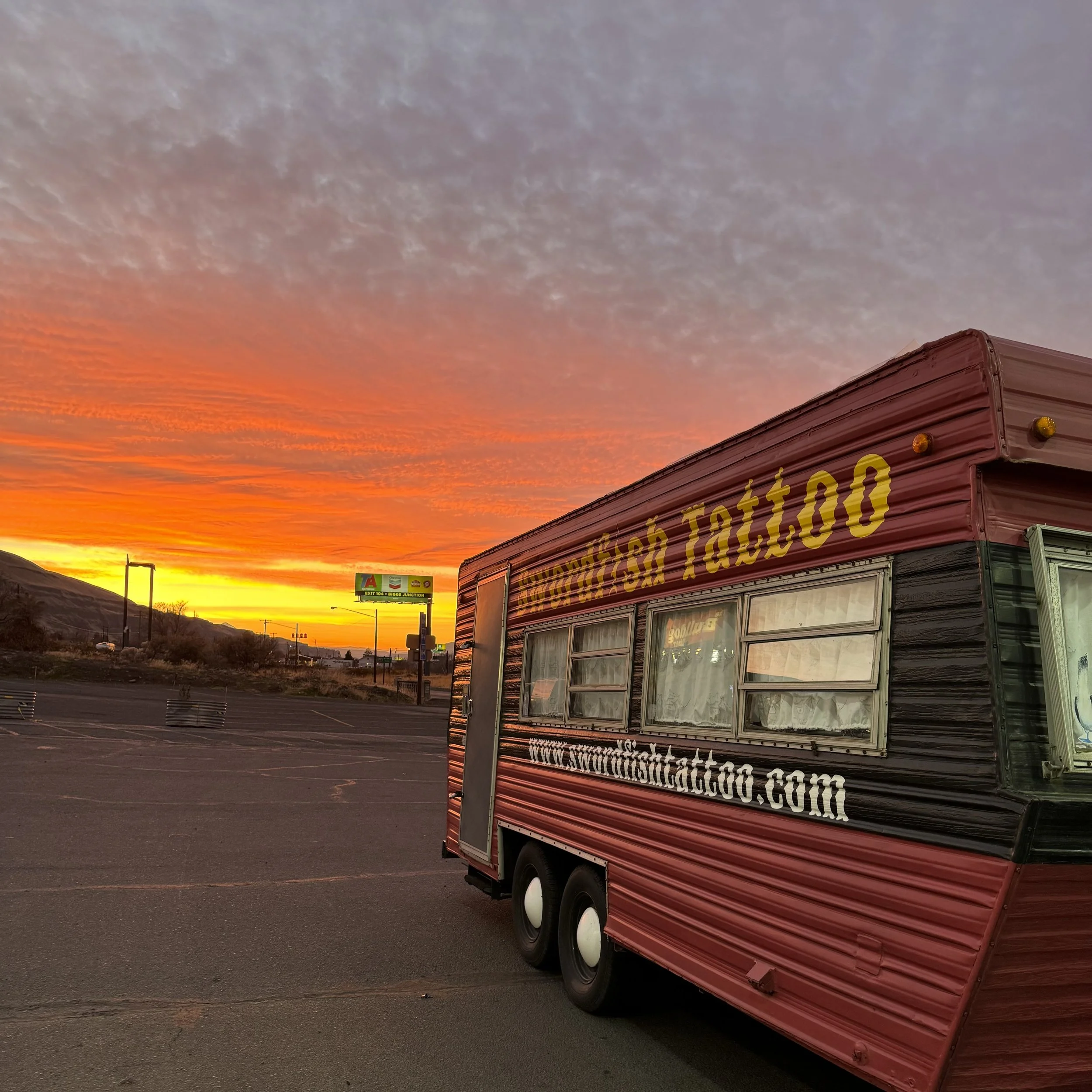 A red food truck with yellow lettering parked in an empty parking lot during a vibrant sunset with orange and purple clouds.