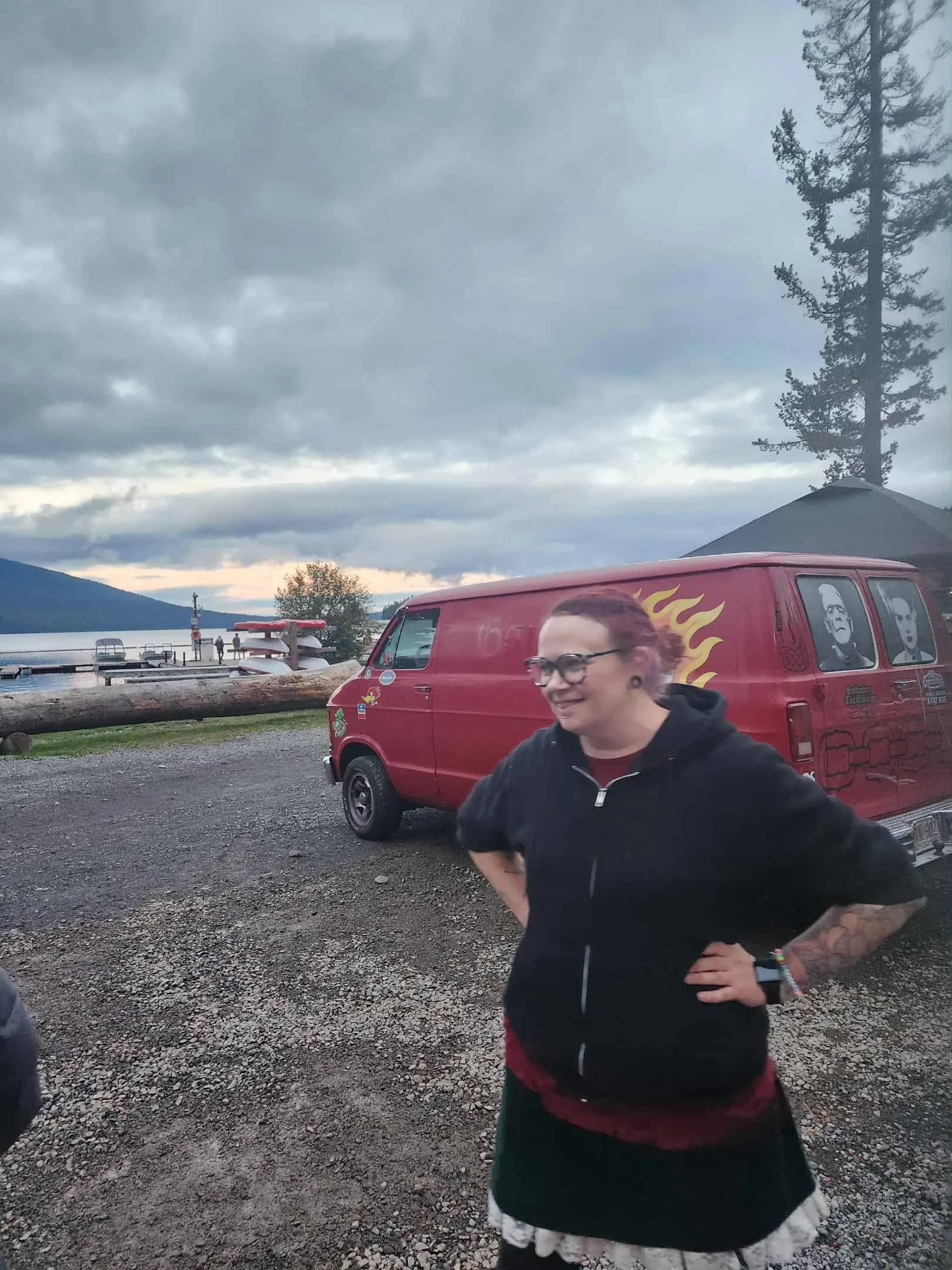 A woman with pink hair and glasses is smiling and standing outdoors near a red van with flame graphics. The background features a lake, mountains, trees, and a cloudy sky.