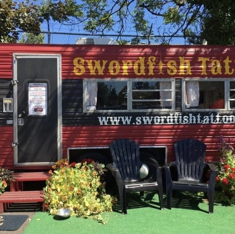 A red and gray food truck with the sign 'Swordfish Tat' and website 'www.swordfishtat.com'. There are two black plastic chairs, potted plants with flowers, and a green outdoor carpet in front of the truck.