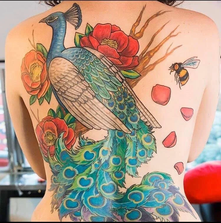 Colorful tattoo of a peacock with blue and green feathers, surrounded by red flowers and a bee flying nearby.