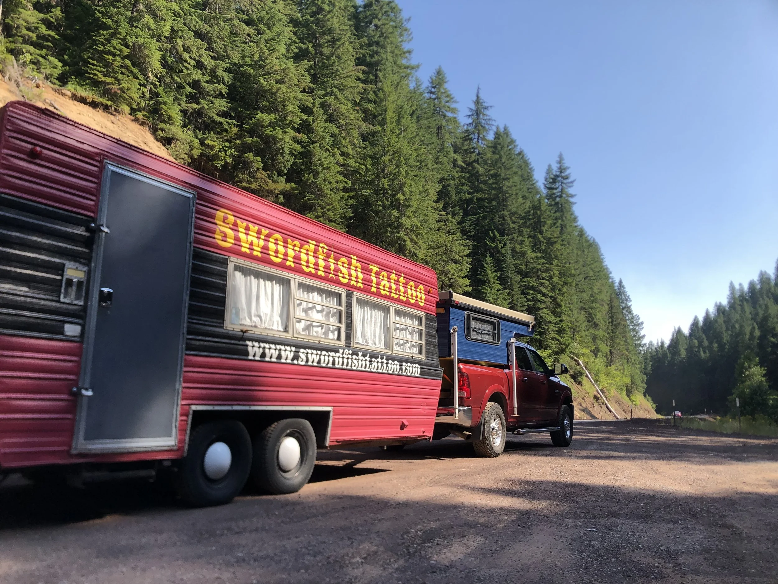 A red truck towing a pink food trailer with yellow lettering that reads 'SWORDFISH TATTOO' and a website URL. The vehicles are parked on a dirt side road surrounded by dense green pine trees under a clear blue sky.