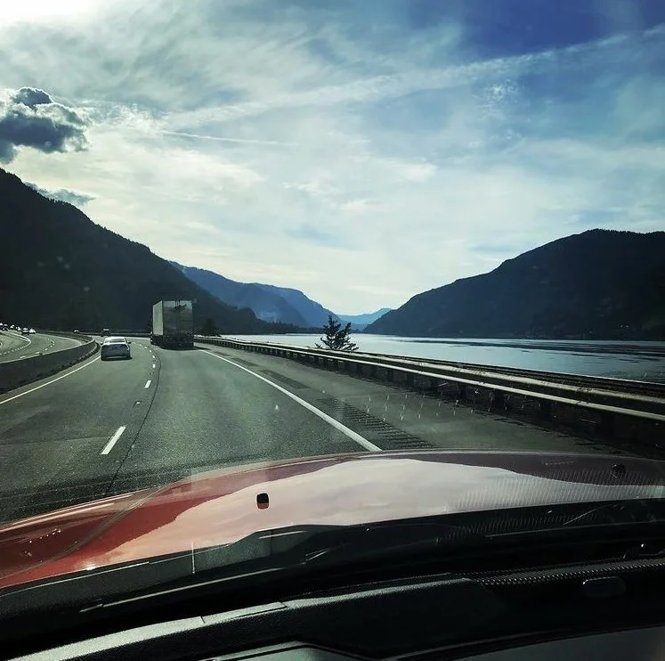 View from inside a car on a highway running alongside a large body of water, surrounded by mountains under a partly cloudy sky.