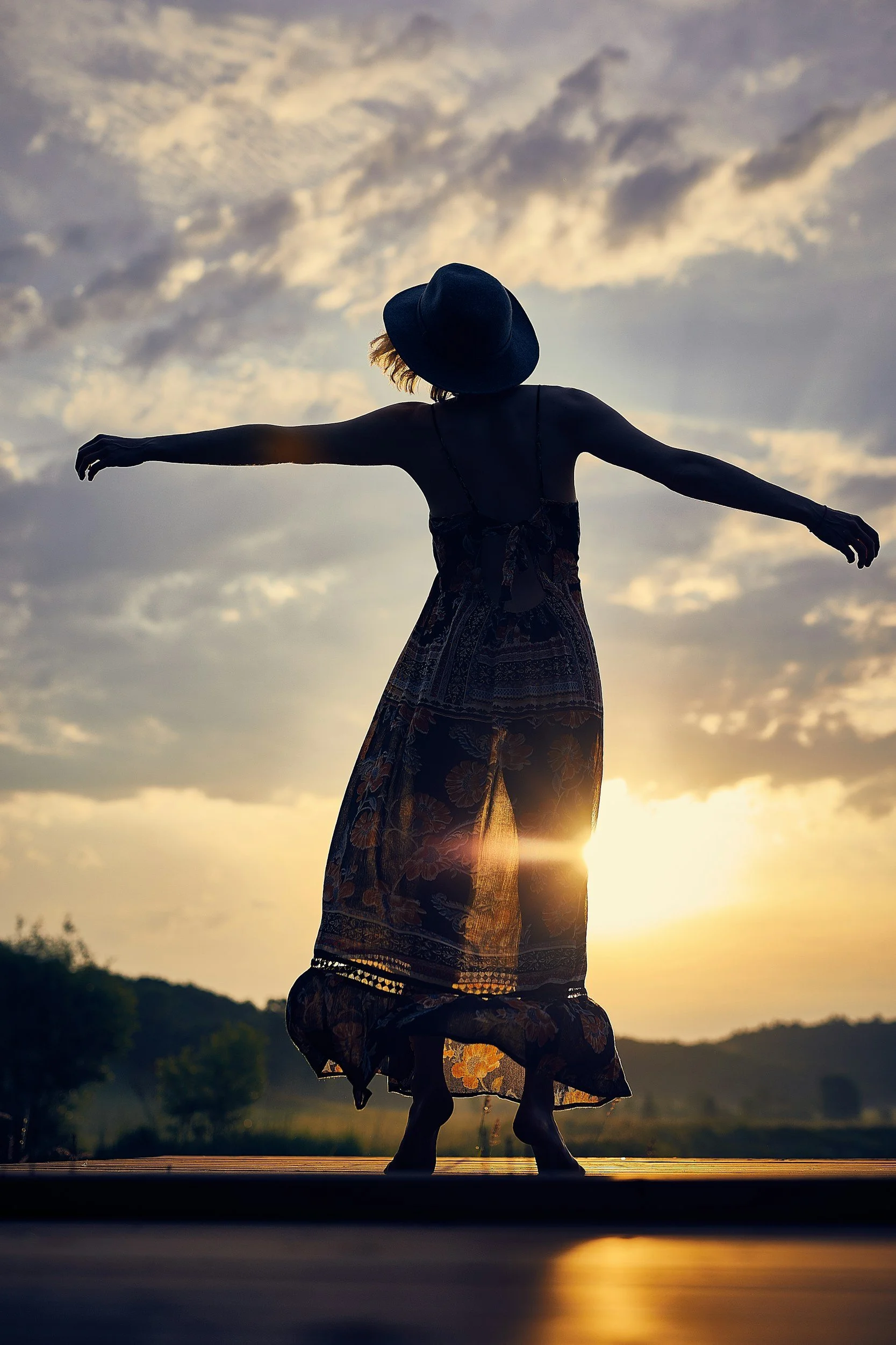 A person balancing on one leg while performing a dance or yoga pose outdoors at sunset, wearing a wide-brimmed hat and flowing clothing, with clouds in the sky.