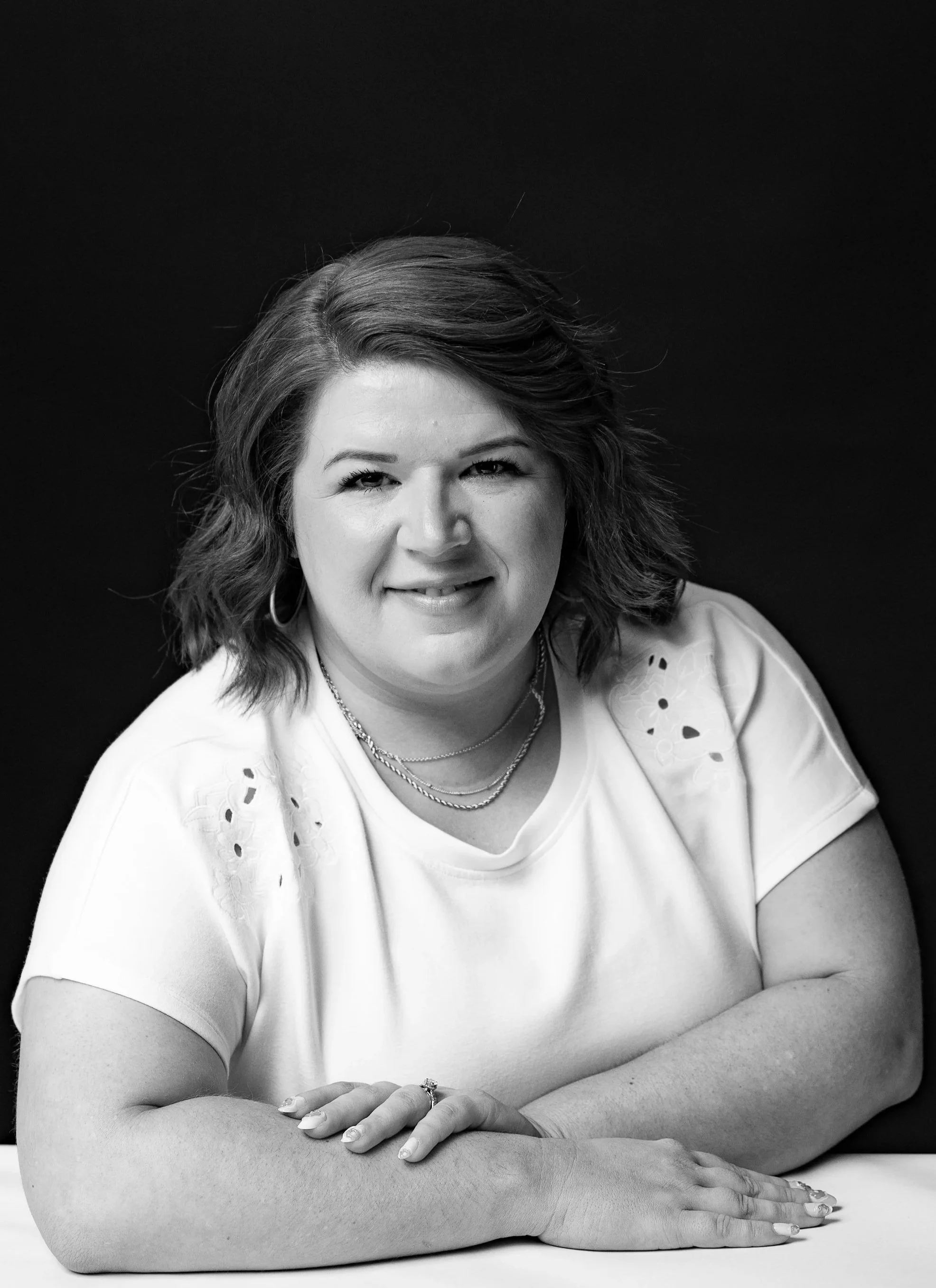 Black and white portrait of a smiling woman with wavy shoulder-length hair, wearing a short-sleeved blouse with eyelet details, layered necklaces, and a ring on her right hand, sitting with her arms crossed on a table against a solid dark background.