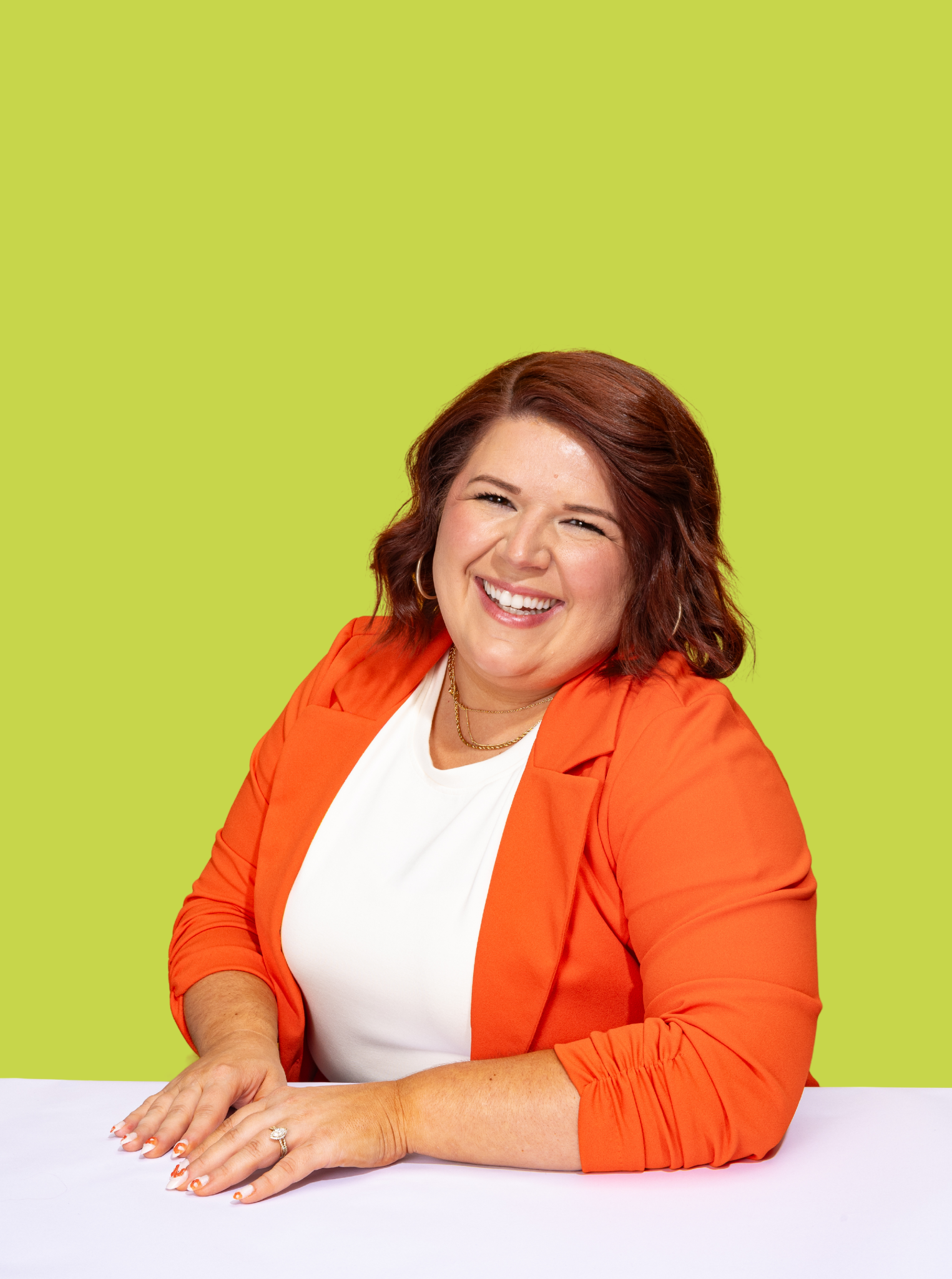 A woman with red hair smiling while sitting at a table against a bright lime green background, wearing an orange blazer and white top.