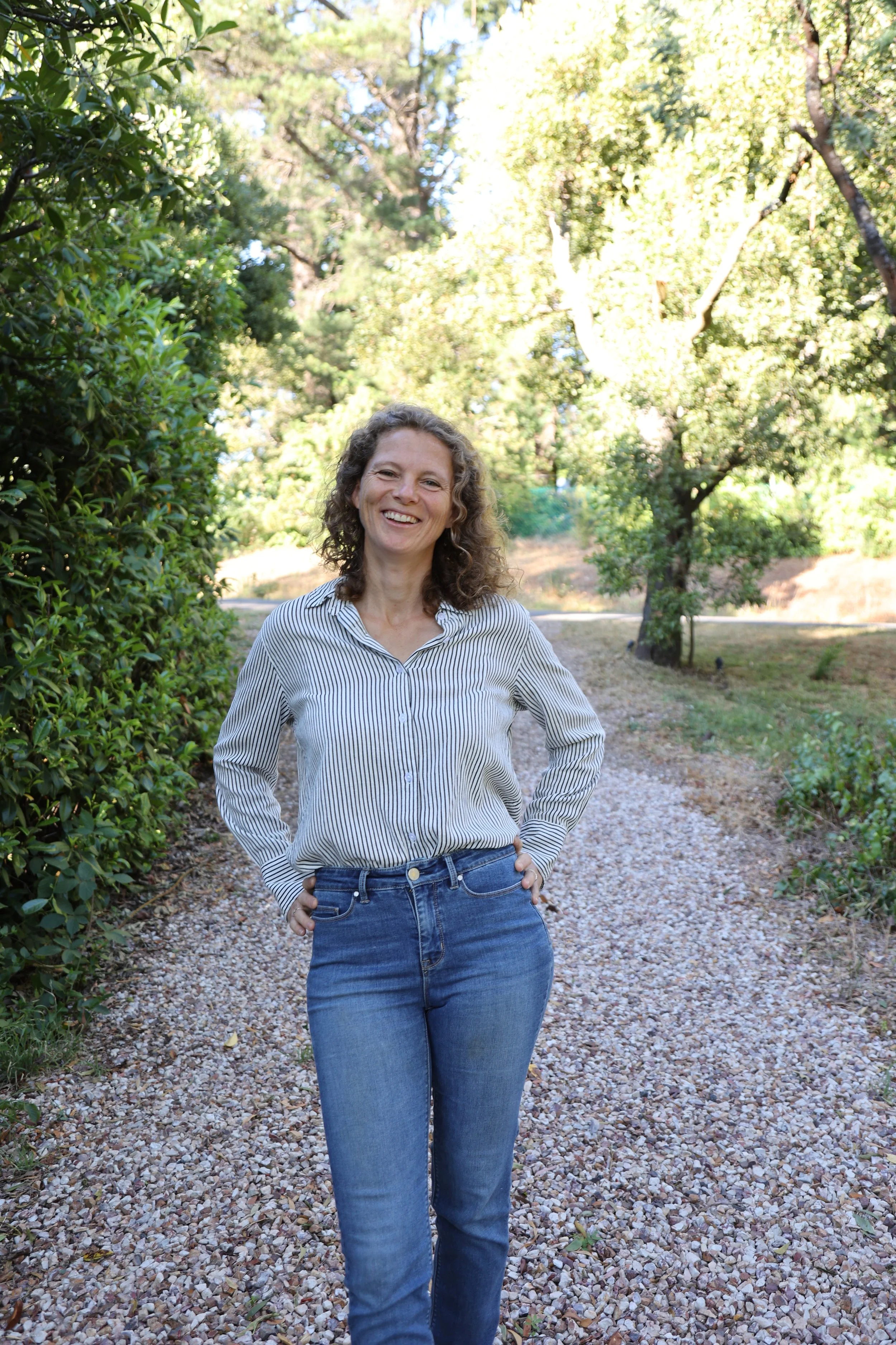 A woman standing outdoors on a gravel path surrounded by green trees, smiling with her hands on her hips, wearing a striped button-up shirt and blue jeans.
