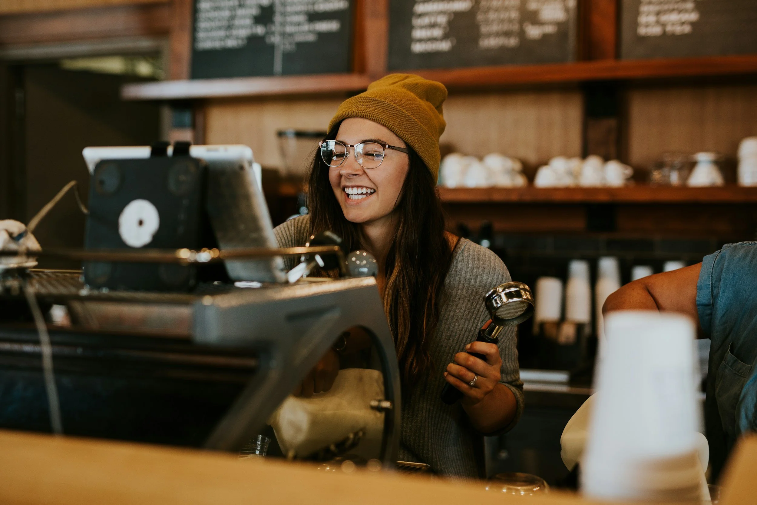 A woman with long dark hair, glasses, and a yellow beanie smiling and holding a microphone in a coffee shop, in front of a coffee counter.