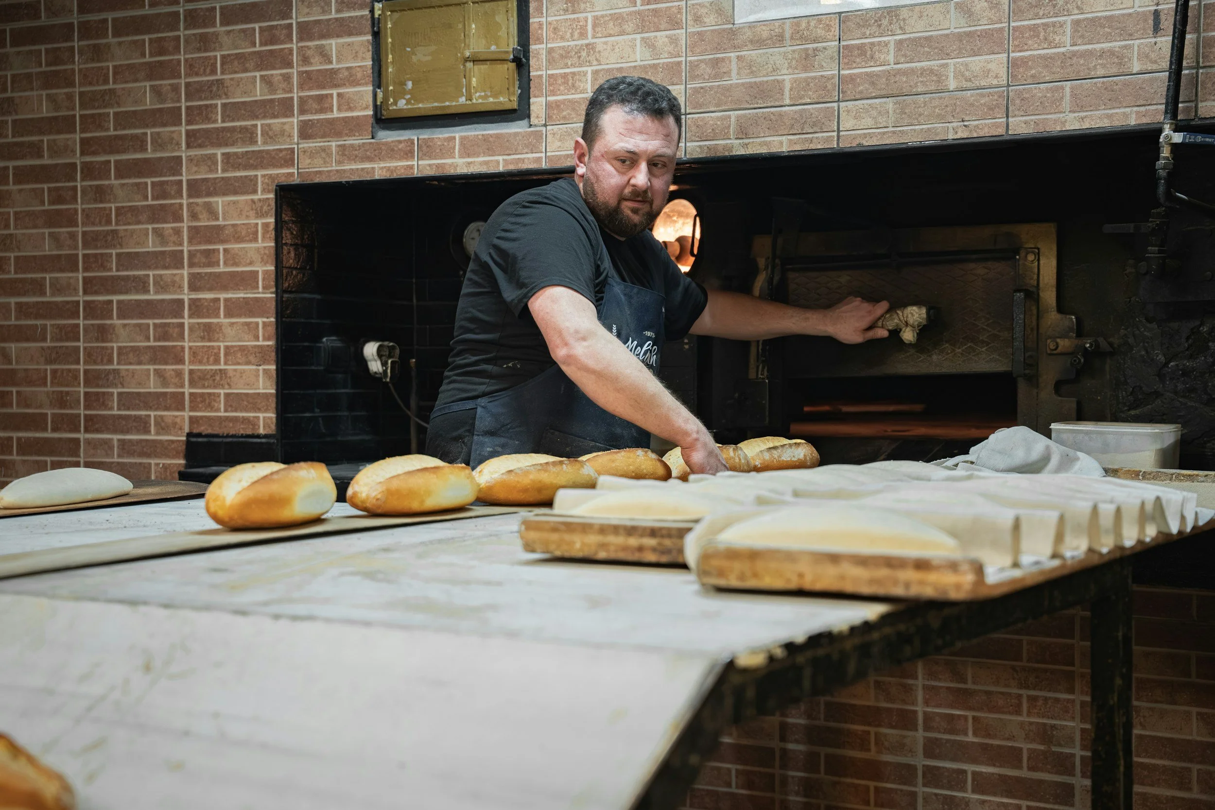 A man with a beard and black t-shirt tending to freshly baked bread loaves in a bakery kitchen with a brick wall background.