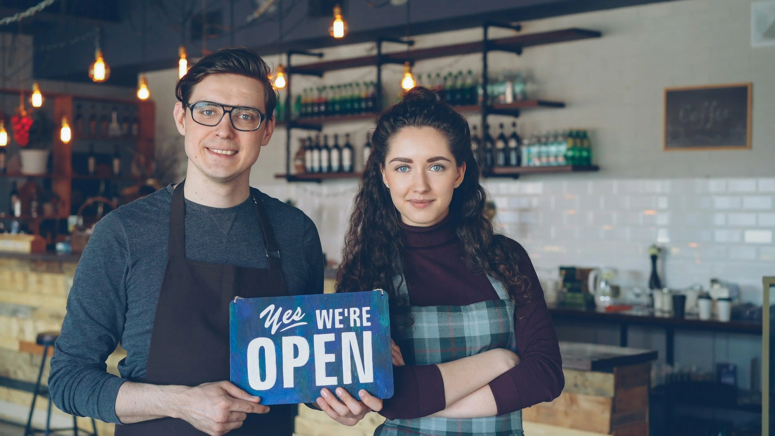 Two people, a man and a woman, standing inside a cafe with warm lighting. The man is holding a blue sign that reads "Yes, we're open" and the woman is standing with her arms crossed. Both are wearing aprons and looking at the camera.