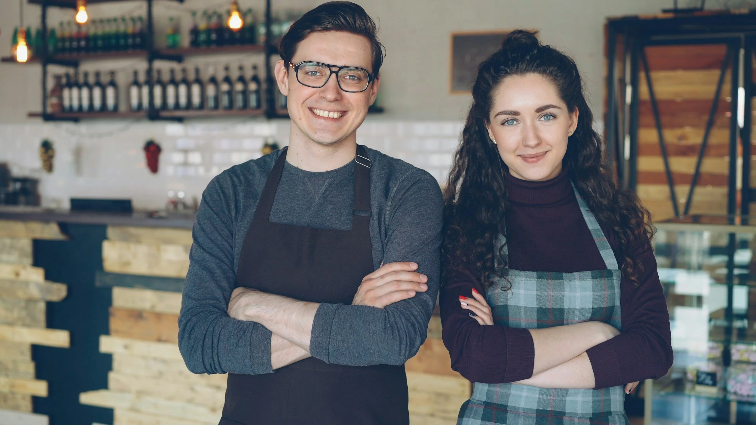 A man and woman standing side by side with arms crossed, inside a cafe or restaurant, smiling at the camera. The man has short brown hair, glasses, and is wearing a dark gray shirt and an apron. The woman has long curly brown hair, blue eyes, and is wearing a dark sweater and a checkered apron.