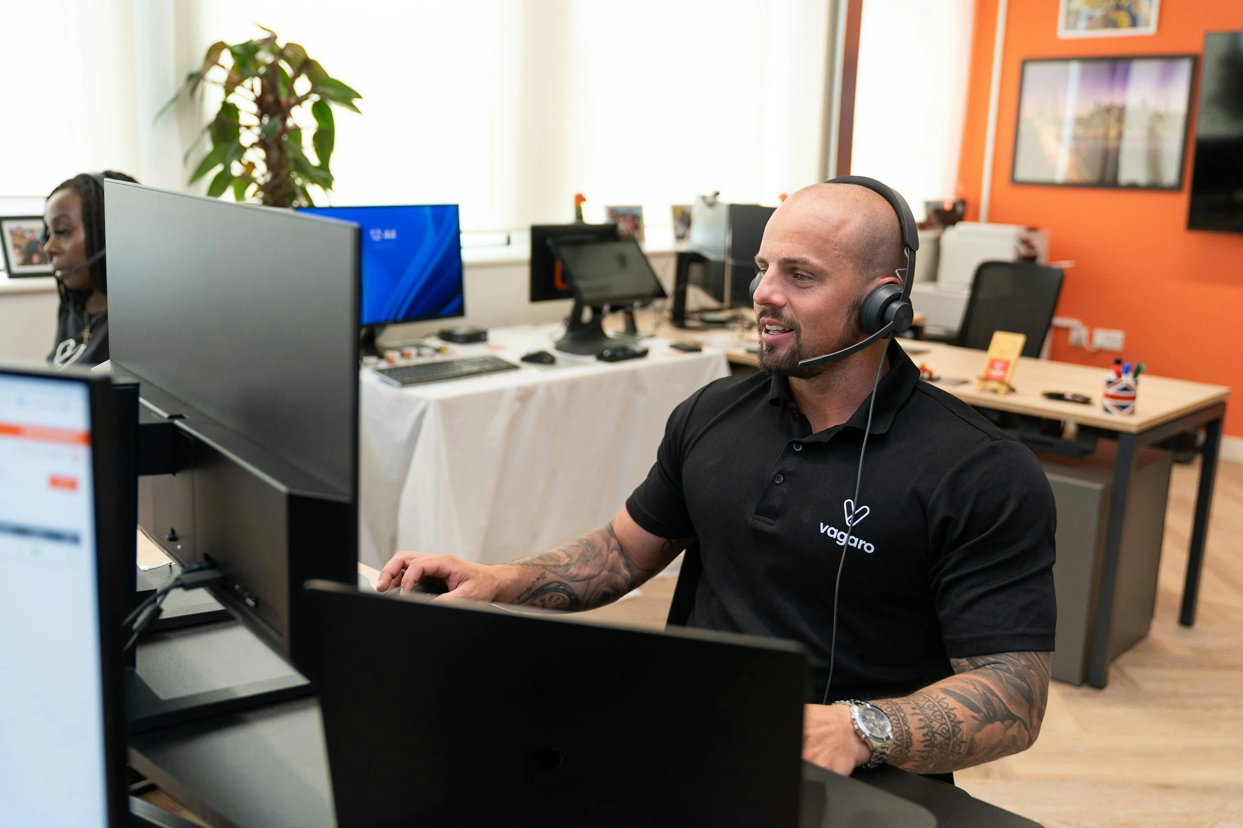 A man with a shaved head, wearing headphones and a black polo shirt with a logo, sitting at a desk in an office, smiling and working on a computer.
