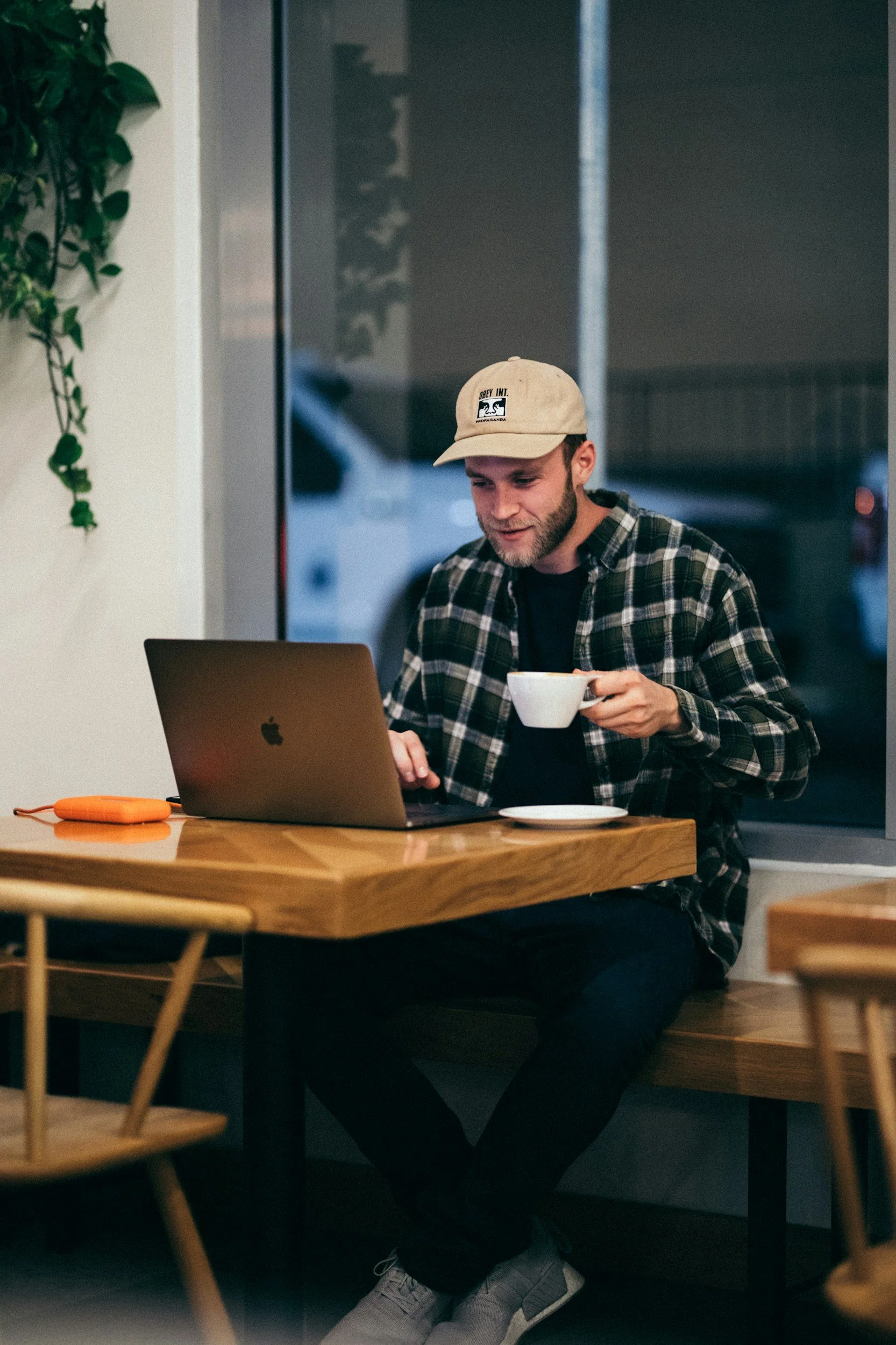 A man wearing a beige baseball cap, a black and white plaid shirt, and black pants sitting on a wooden bench in a cafe or restaurant, holding a white cup, working on a silver laptop.