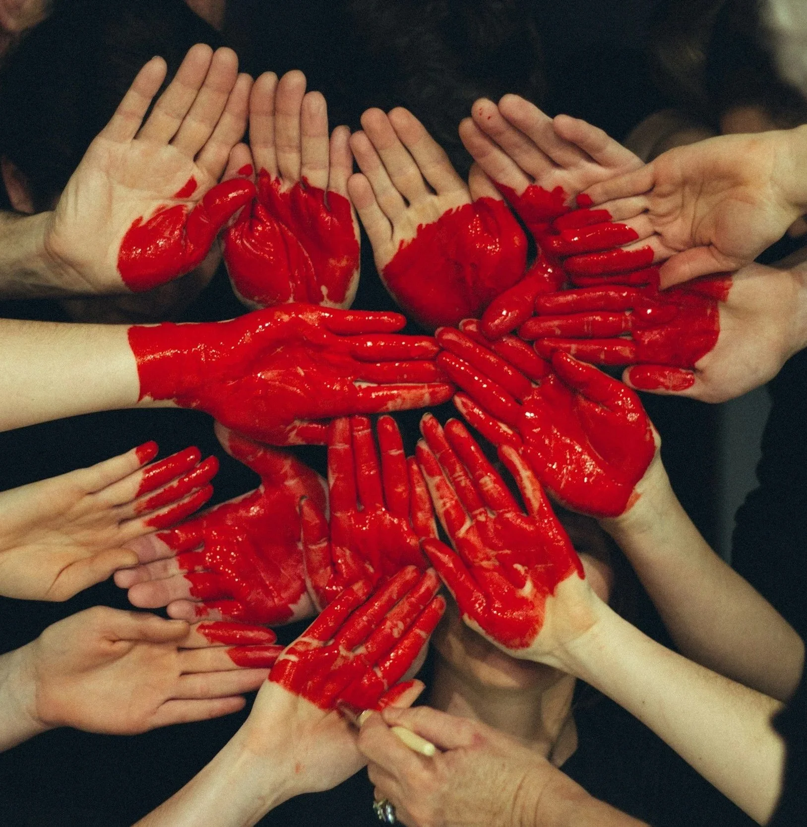 Multiple hands covered in red paint, with some hands forming heart shapes, others with fingers extended, against a dark background.