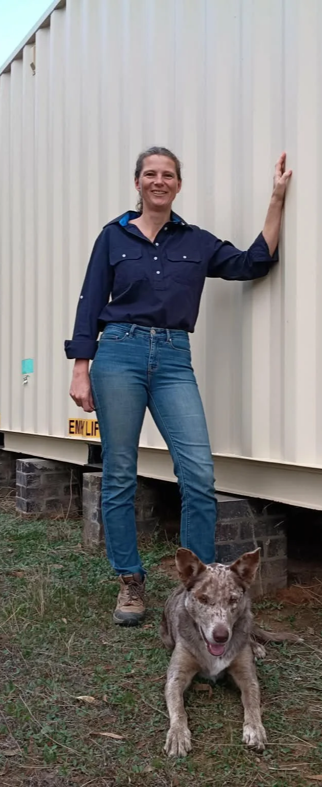 A woman standing outdoors next to a beige metal building, smiling, with her hand resting on the wall. A dog with a speckled coat of white and brown fur is sitting on the grass in front of her.