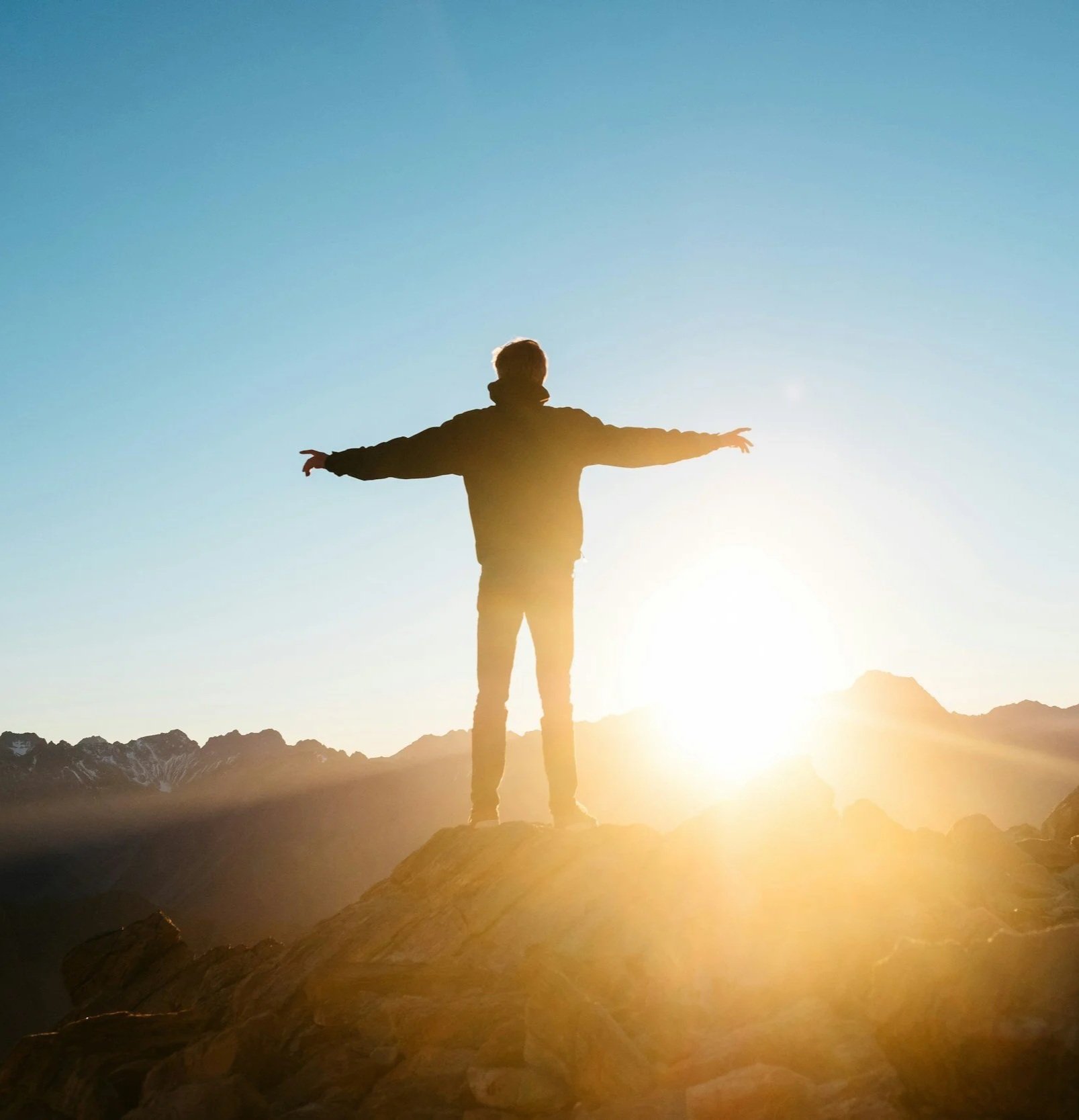 Person standing on rocky terrain with arms outstretched in front of a bright sunrise or sunset over mountains.