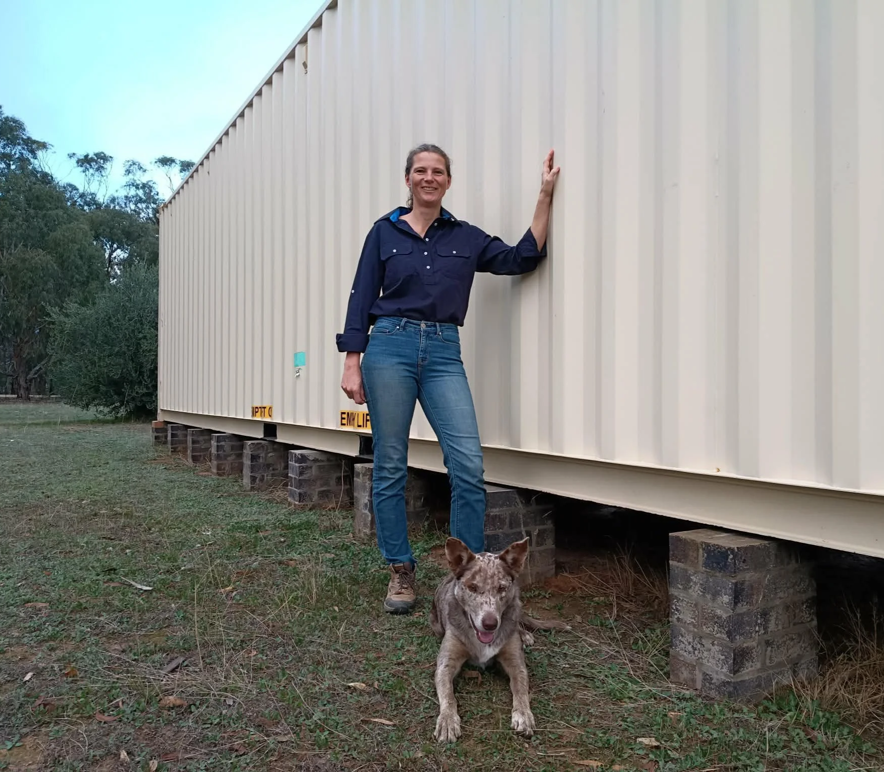A woman with long brown hair wearing a navy blue shirt and jeans standing outdoors next to a large beige shipping container with a dog sitting in front of her.