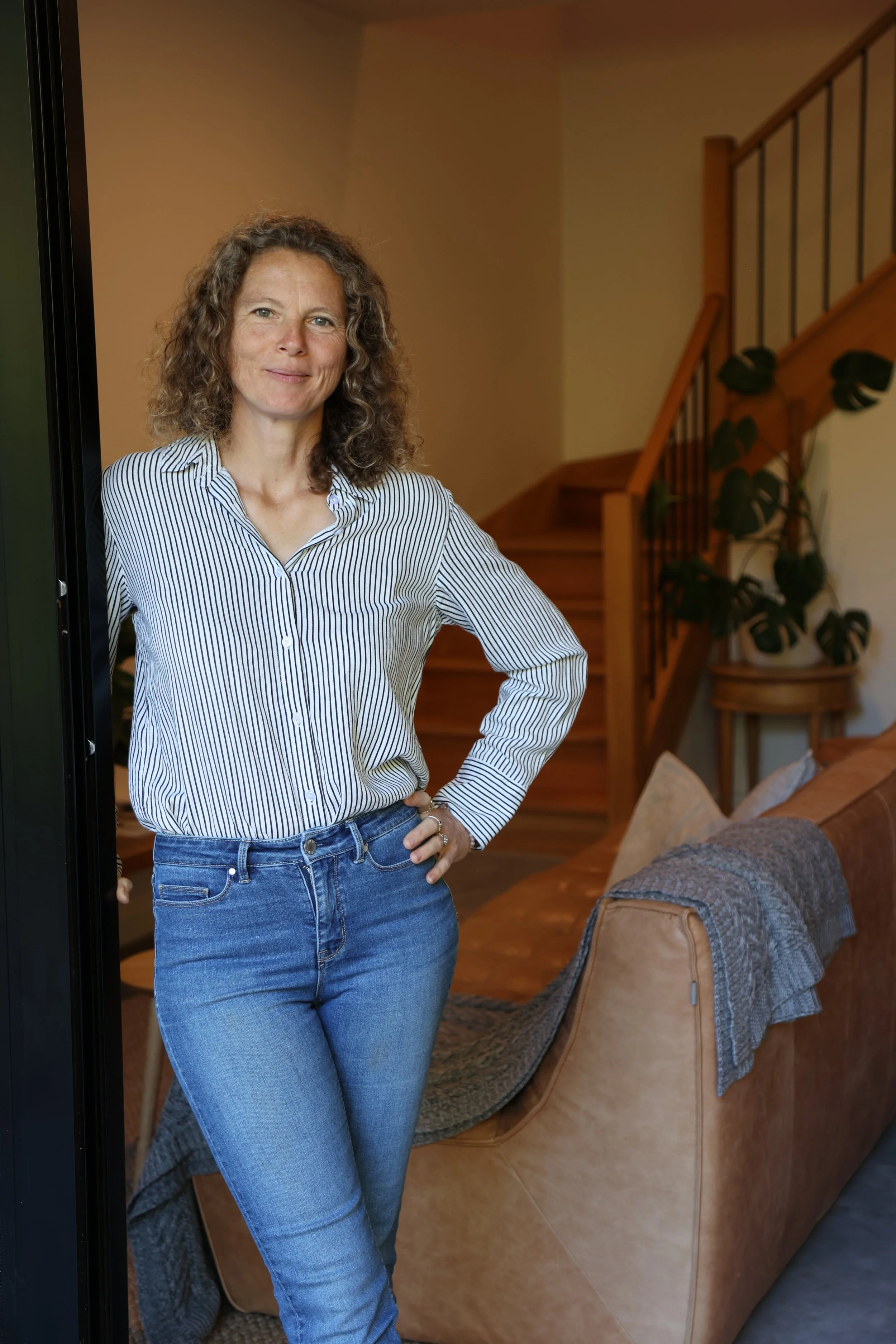 A woman with curly hair wearing a striped shirt and jeans standing indoors near a staircase and a brown armchair.