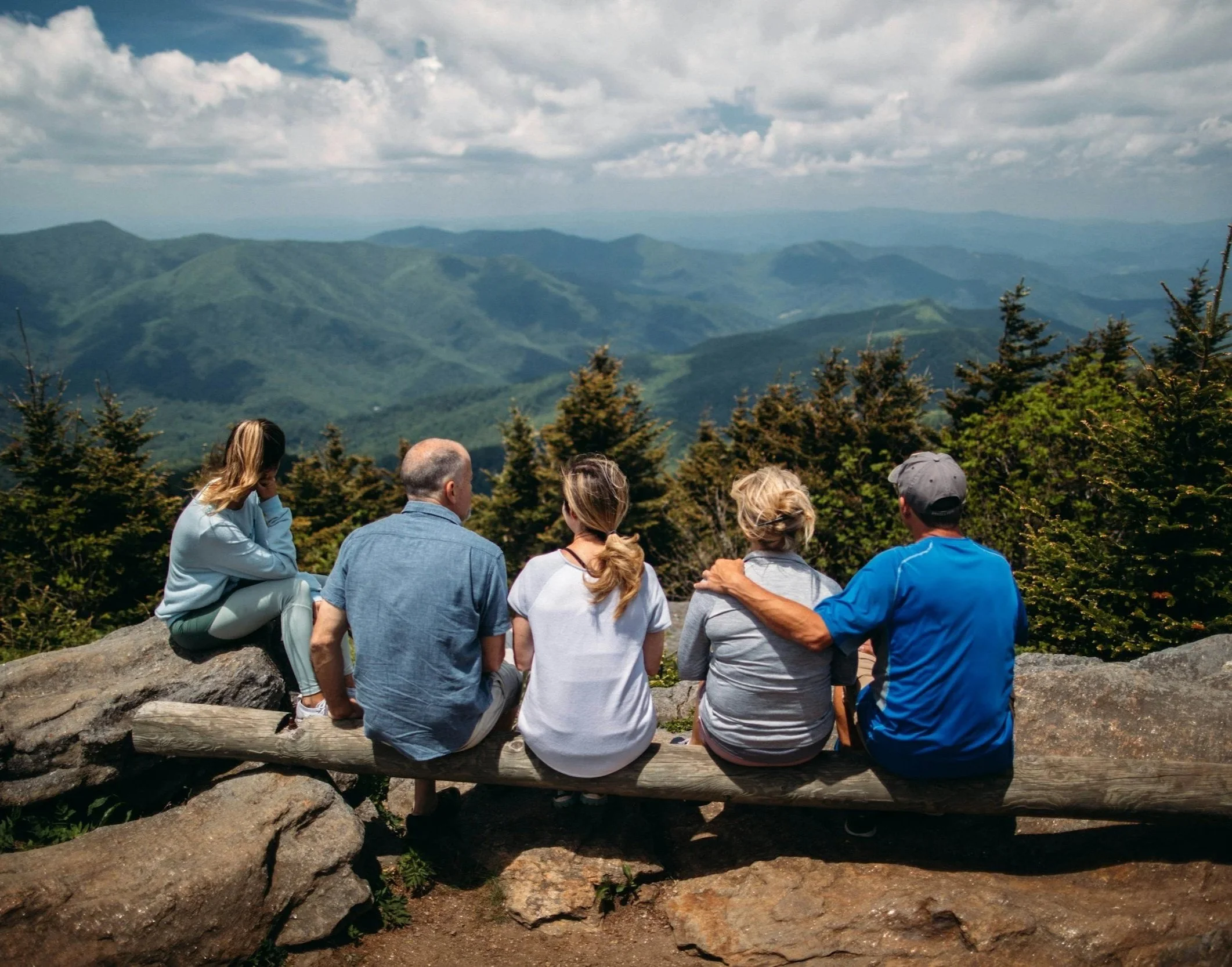 Group of people sitting on a log and looking out at a mountain landscape with clouds in the sky.