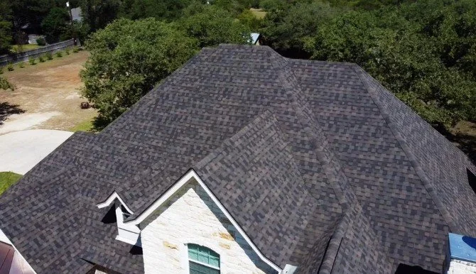 A large house roof with multiple gables and dark asphalt shingles, surrounded by trees and neighboring properties.