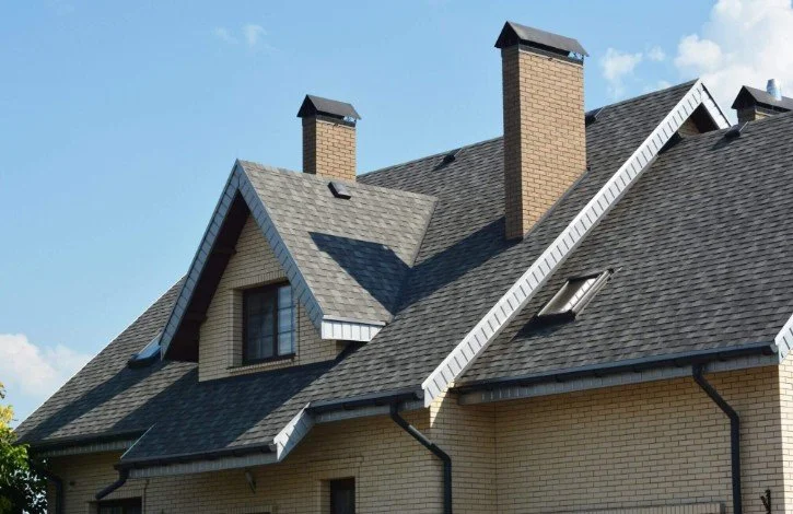 Close-up of a house rooftop with brick chimneys, dormer window, gray shingles, and gutter system, under a partly cloudy sky.
