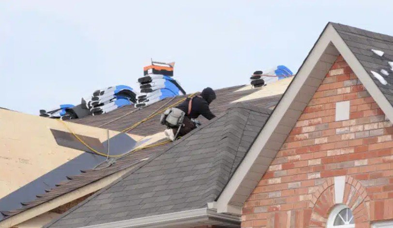 A construction worker on a roof installing shingles, with building materials and tools visible in the background.